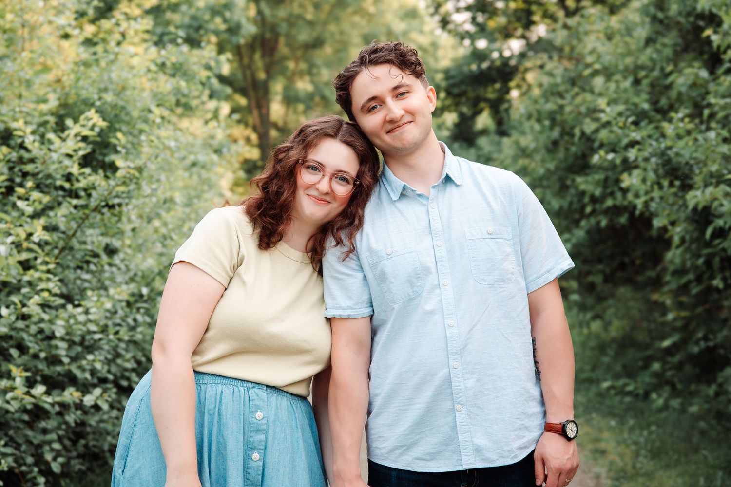 Young couple smiling together outdoors on a sunny day surrounded by lush green trees and foliage.