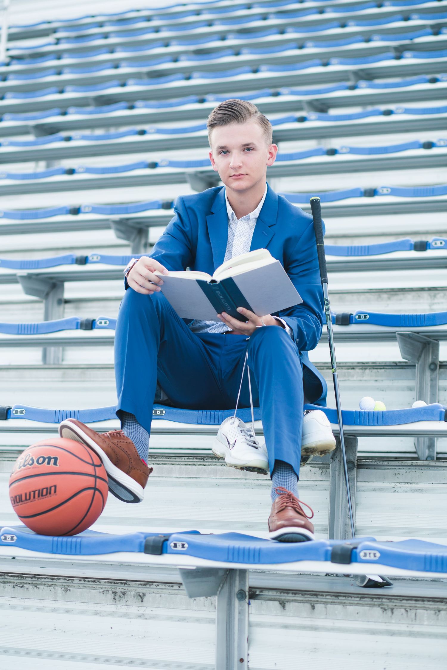 Young man in blue suit reading a book in stadium bleachers with a Wilson basketball nearby.