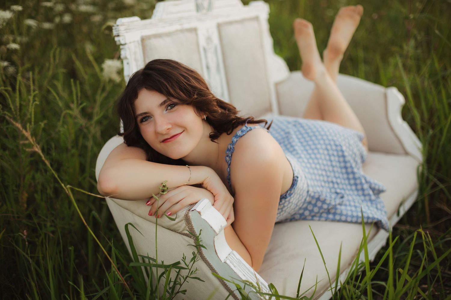 Young woman in blue dress lying on white vintage chair in green meadow, smiling at camera.