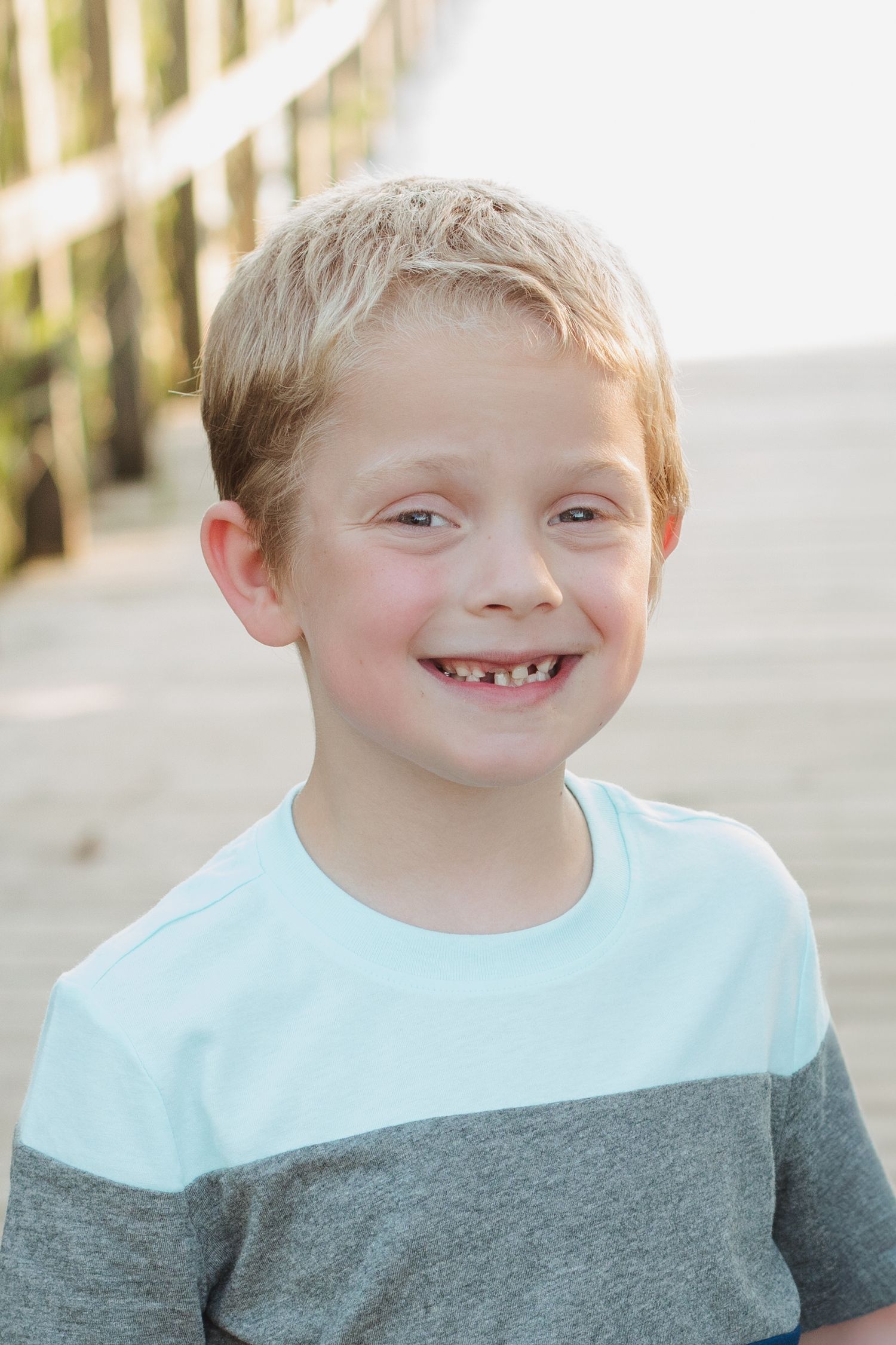 Happy young boy with missing front teeth grins widely on bright sunny wooden boardwalk.