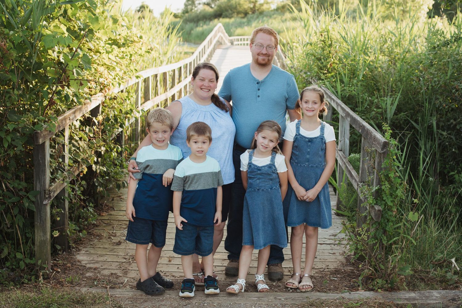 A family of six smiles together on a wooden bridge surrounded by green foliage.