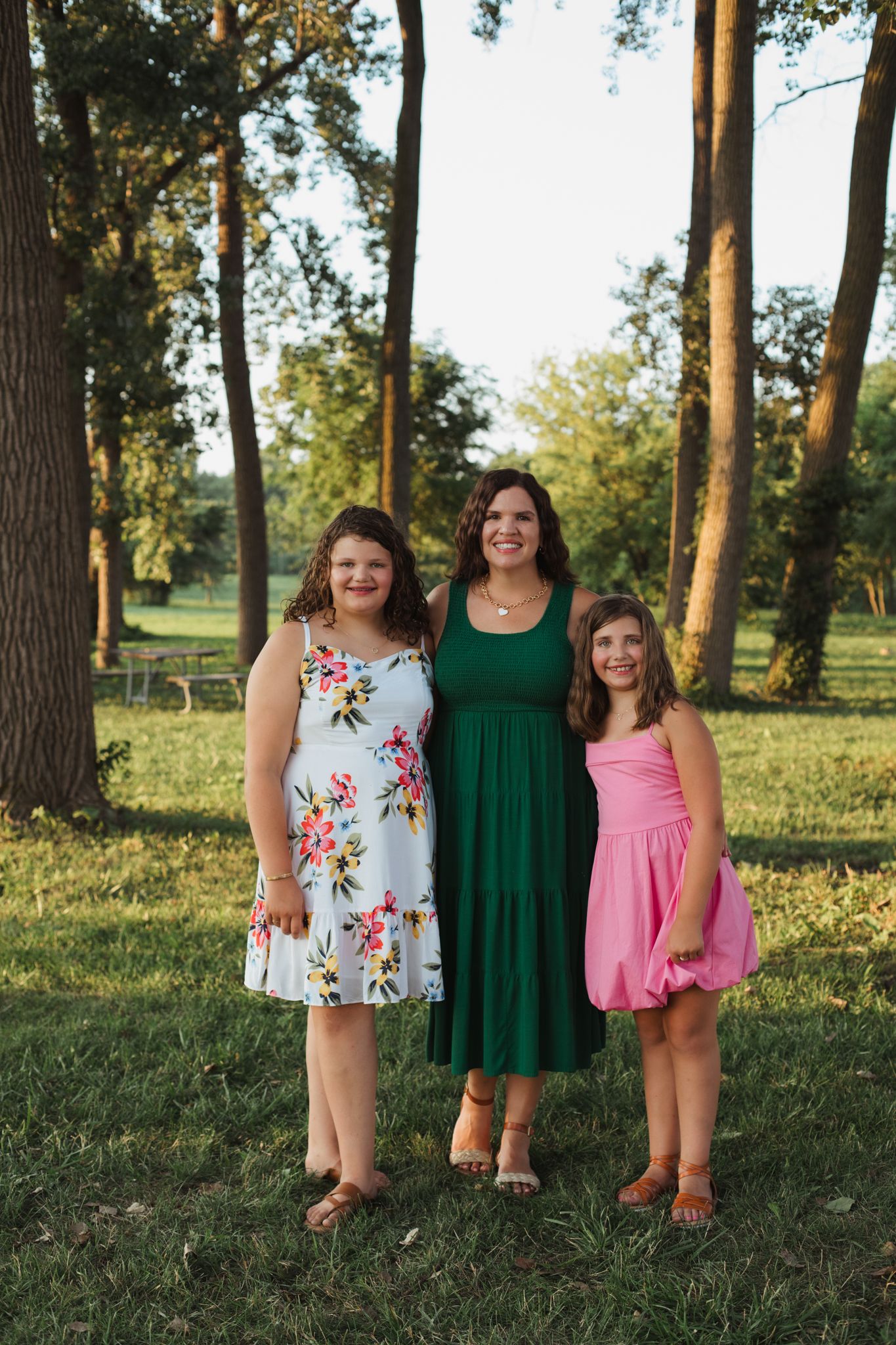 Woman in green dress poses with two girls in floral and pink dresses outdoors.