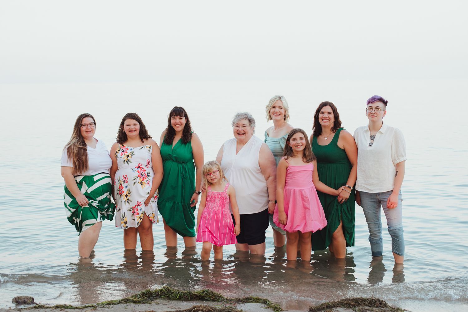 Group of women and girls posing together on rocky shoreline near calm water for a family beach photo session.