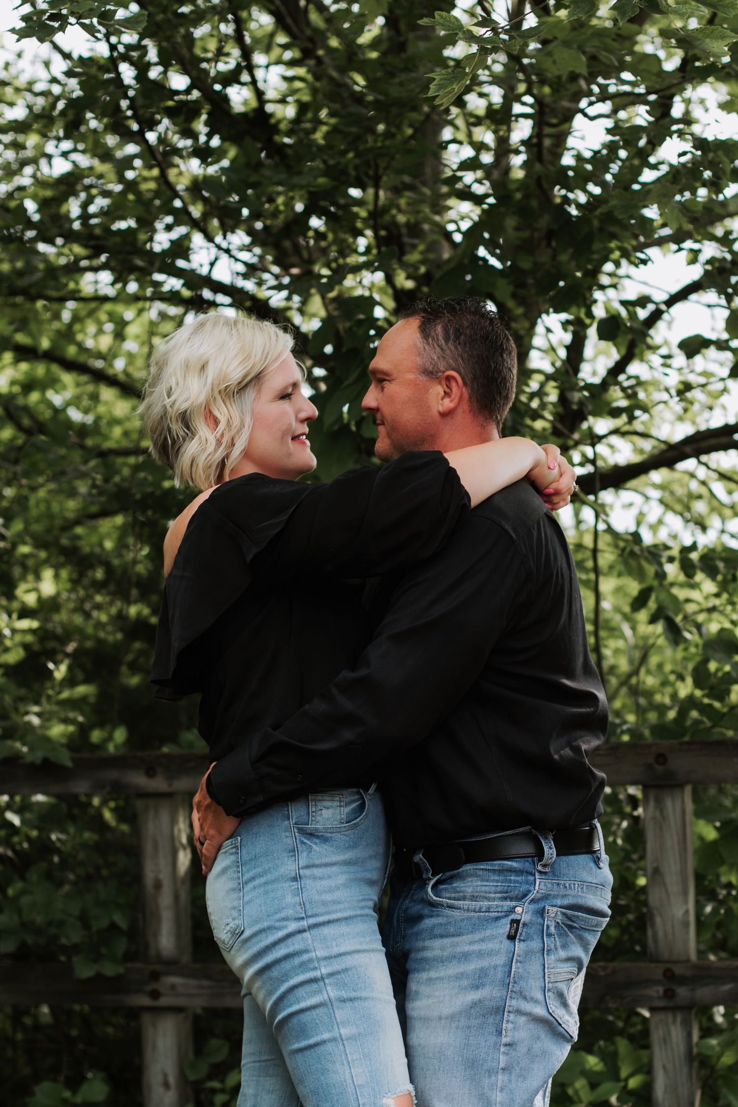 Couple embracing and gazing at each other lovingly near a wooden fence outdoors.