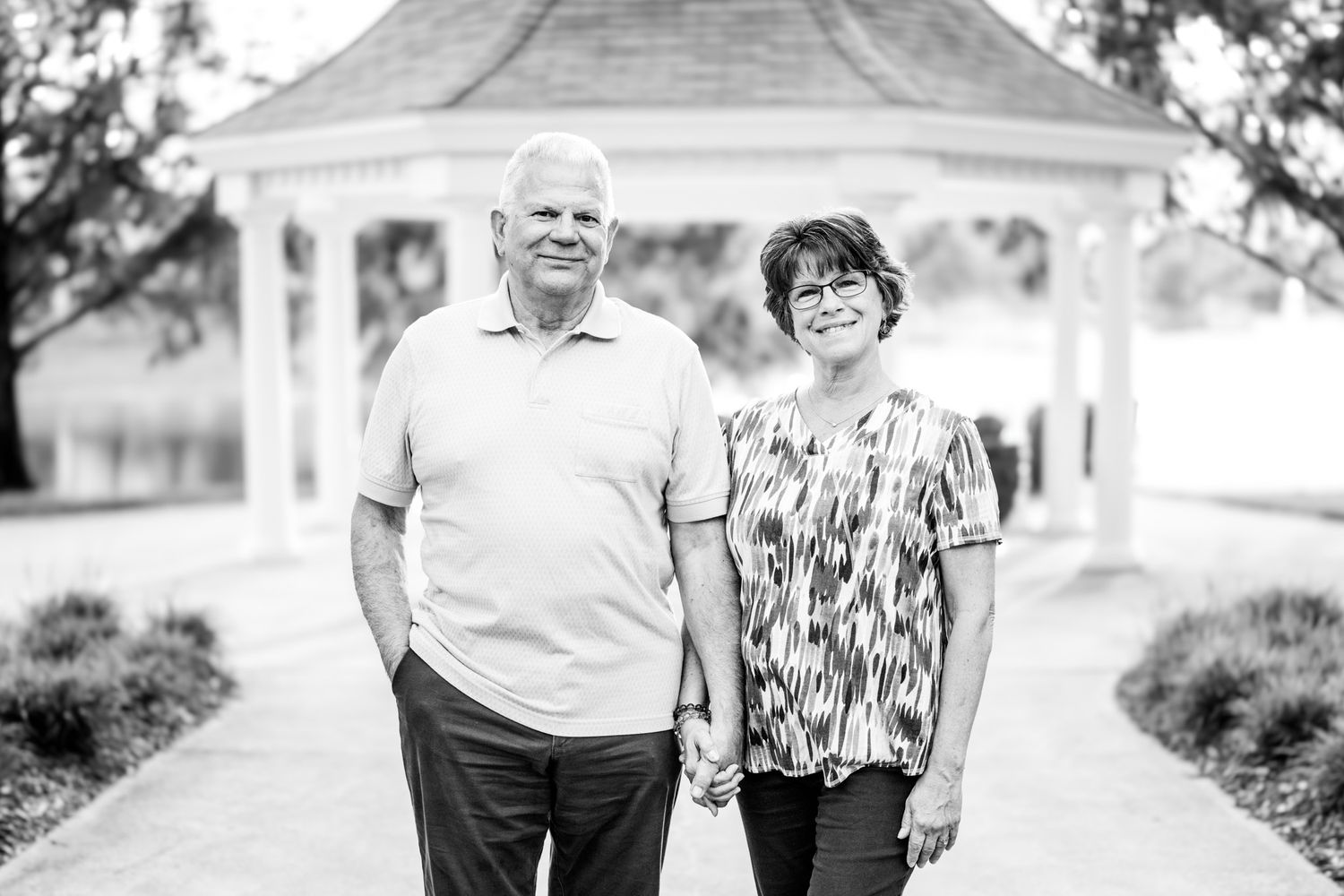 Black and white photo of a smiling couple holding hands in front of a gazebo outdoors.