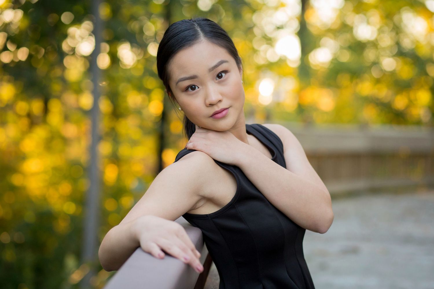 Young Asian woman in black dress leaning on railing in autumn park with golden foliage background.