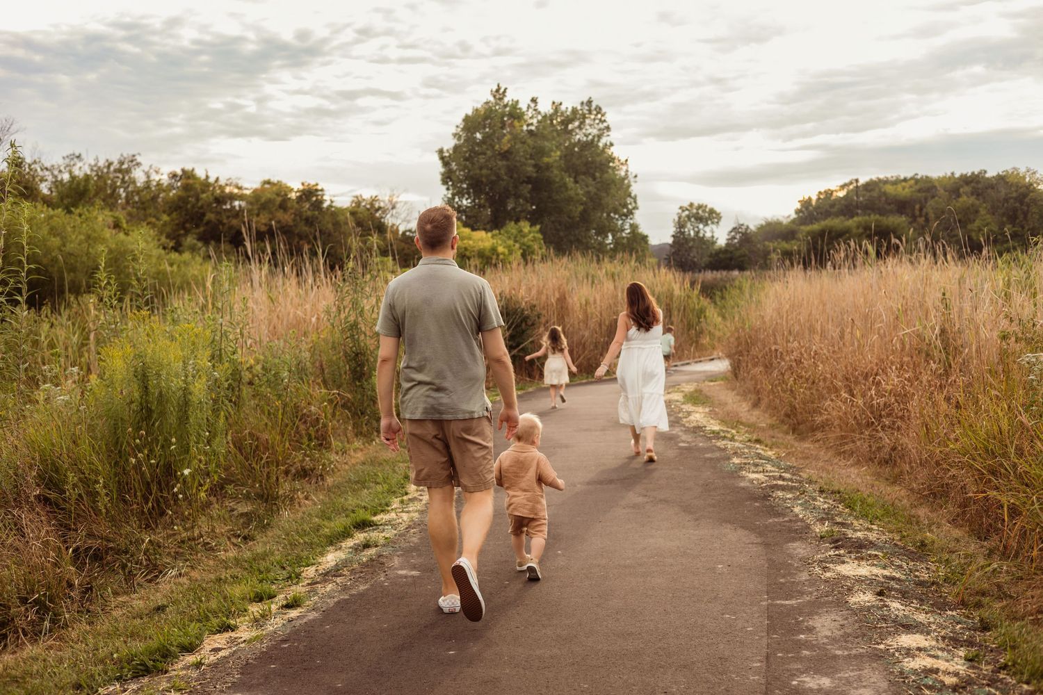 Family of four walking away on a scenic path surrounded by tall grass and trees.