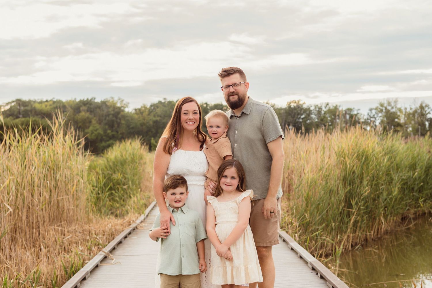 Family of five posing on a wooden boardwalk surrounded by tall grass and greenery on a cloudy day.