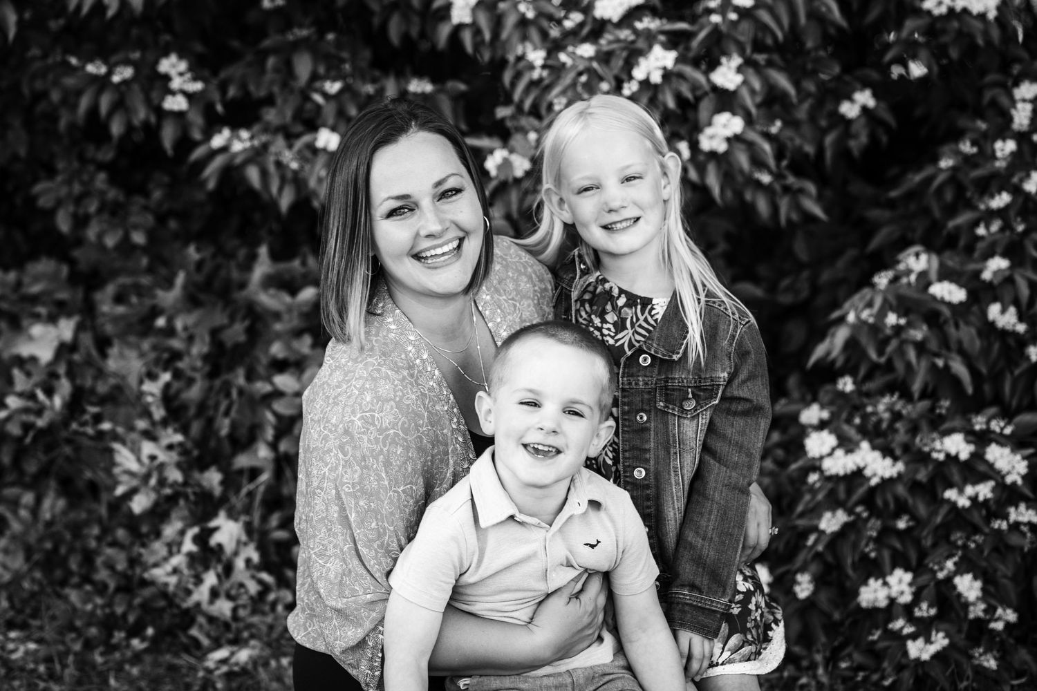 Black and white photo of a smiling mother with two young children outdoors near flowering bushes.