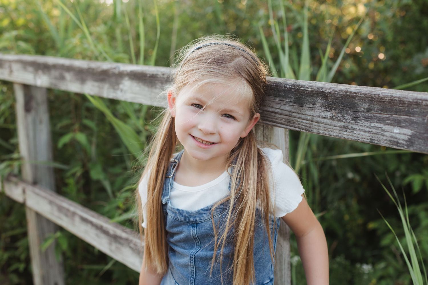 Adorable young girl in denim overalls smiling while leaning against a wooden fence.
