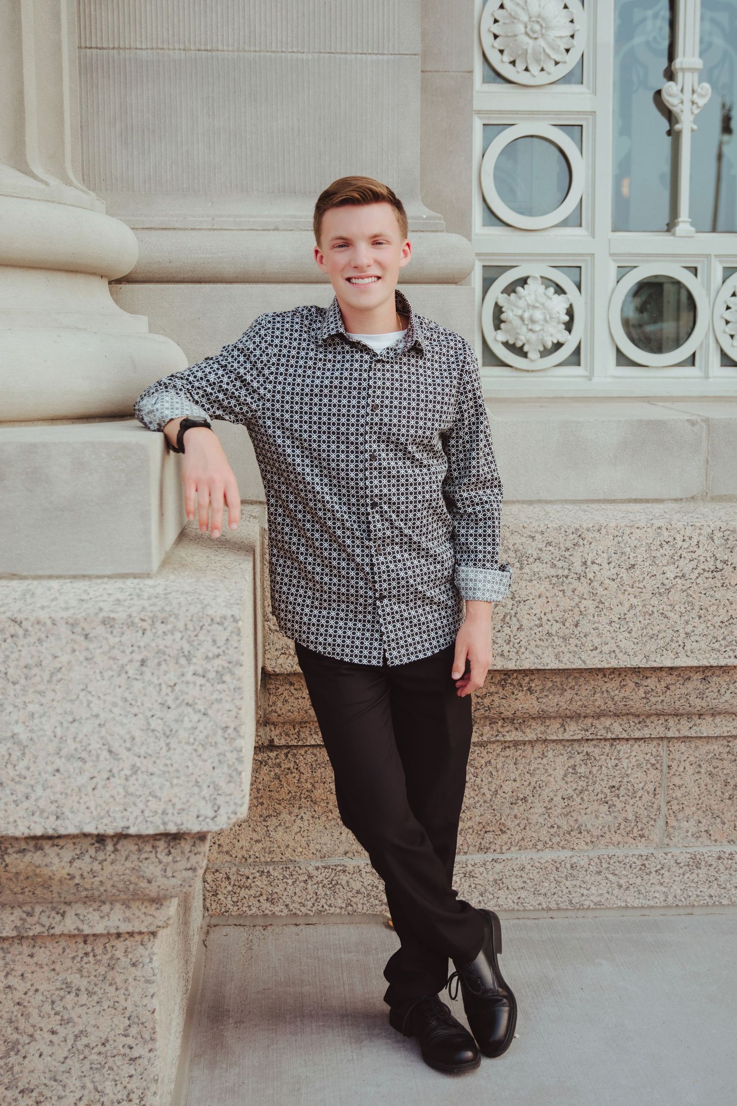 Smiling young man in patterned shirt leaning against stone column outdoors.