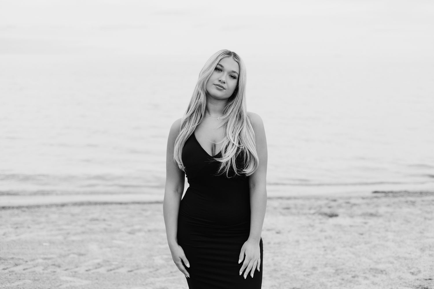 Blonde woman in fitted black dress posing confidently on sandy beach in black and white photo.