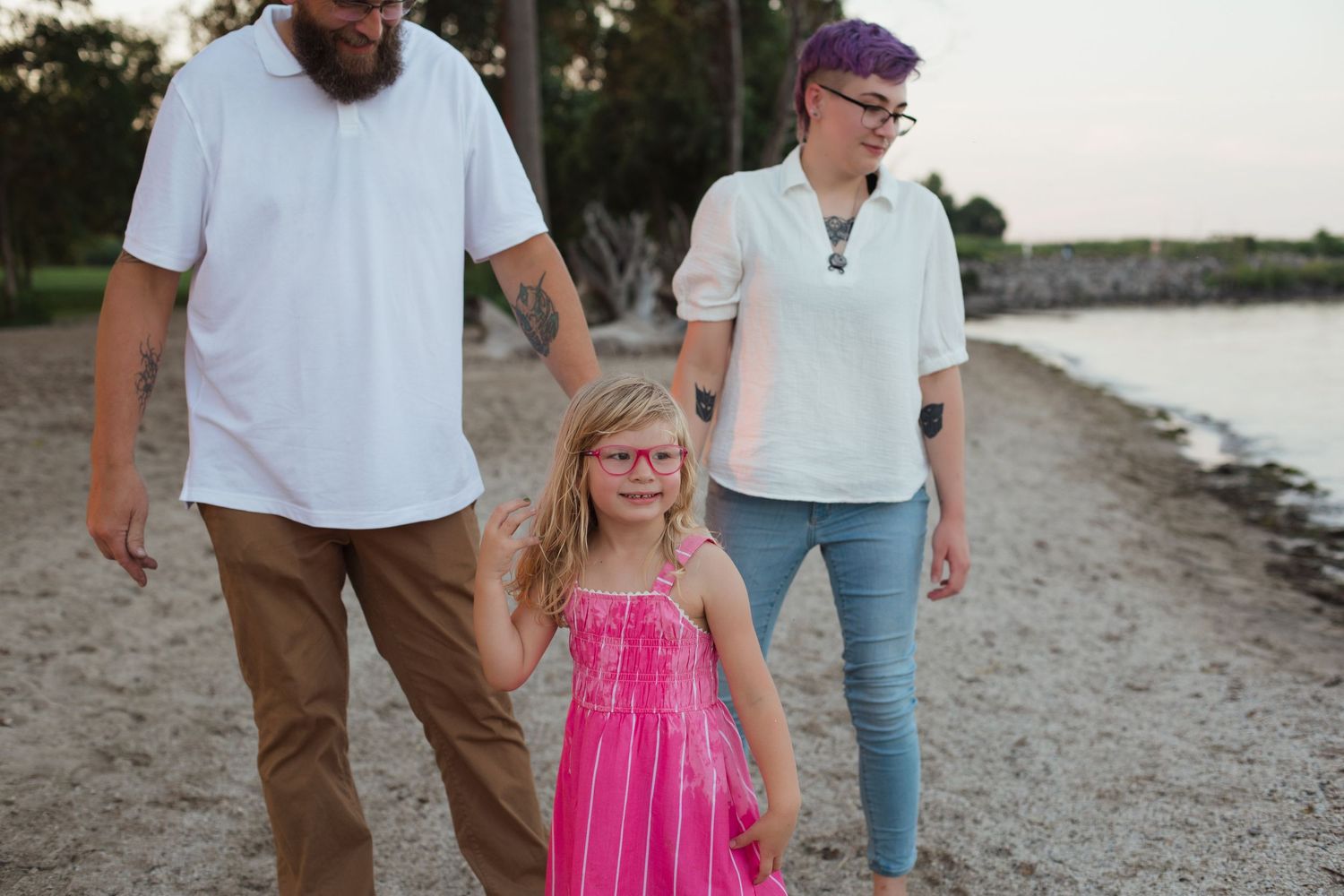 Family members walk along a beach shoreline at sunset wearing casual attire.