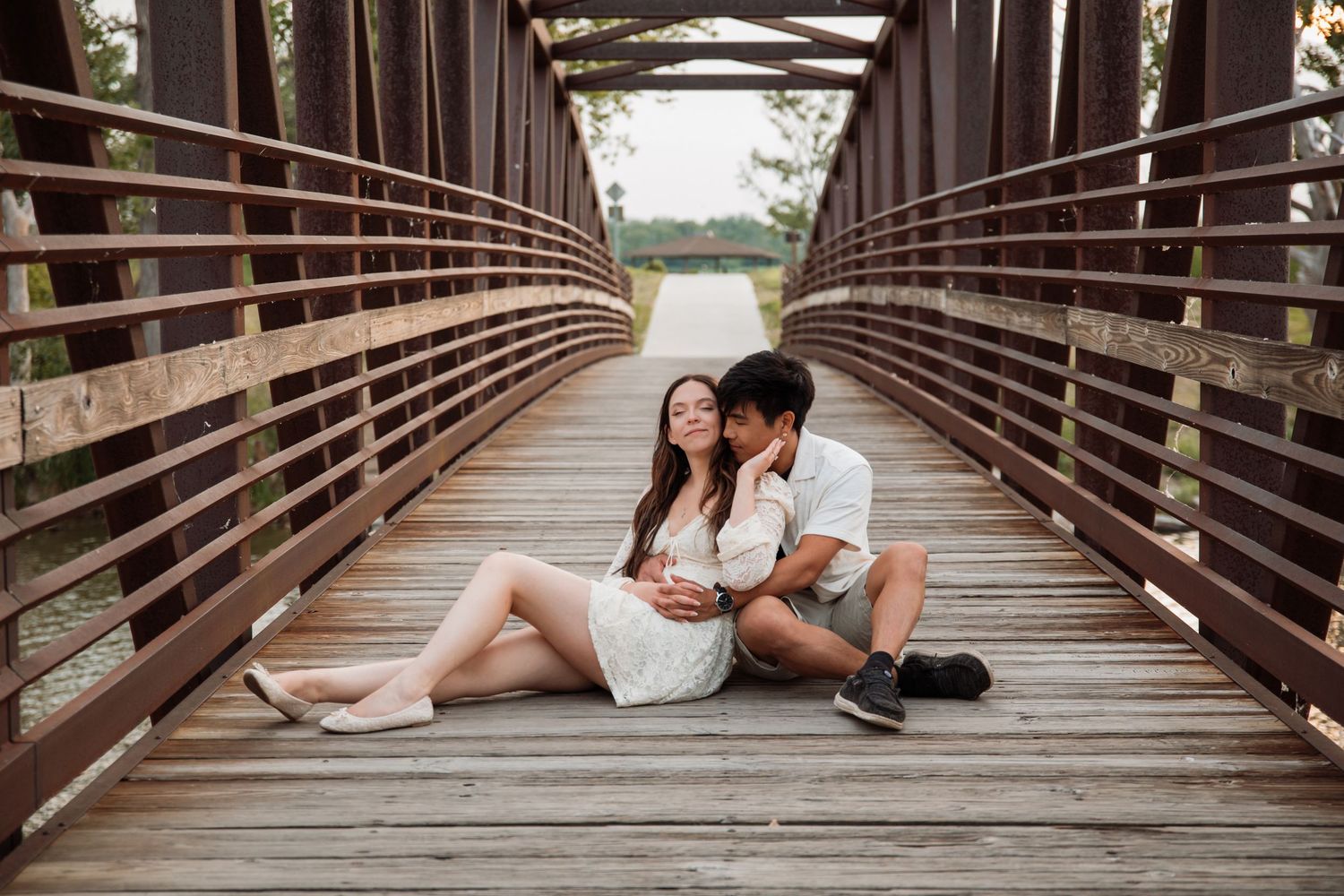 Couple sitting together on a wooden bridge, sharing a tender moment in an outdoor park setting.
