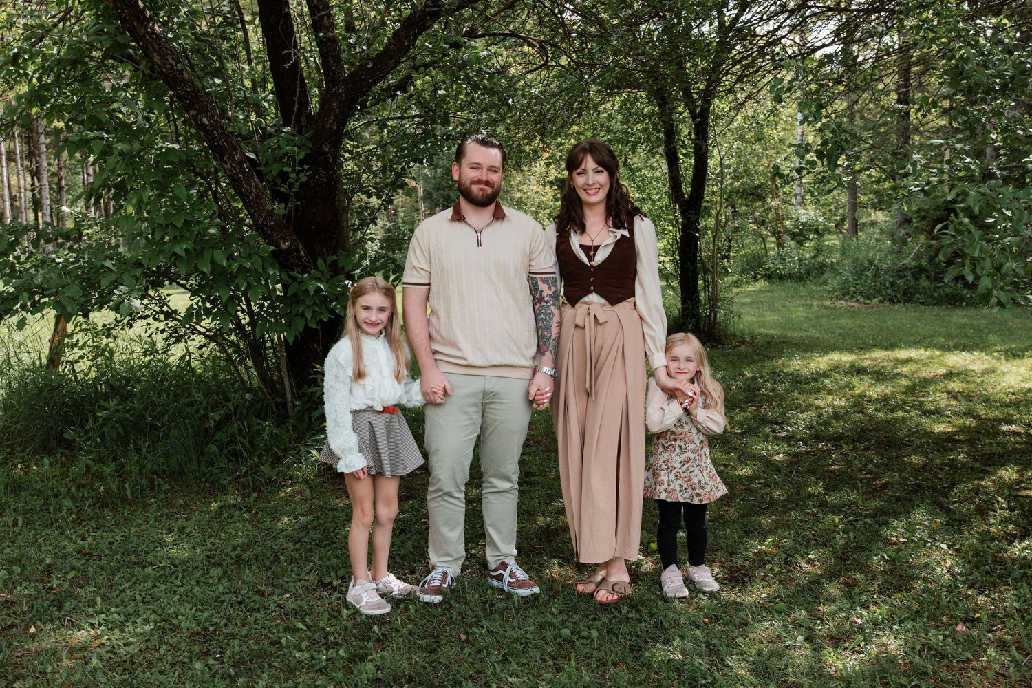 Family of four posing outdoors in a lush green garden, parents with two young daughters smiling together.