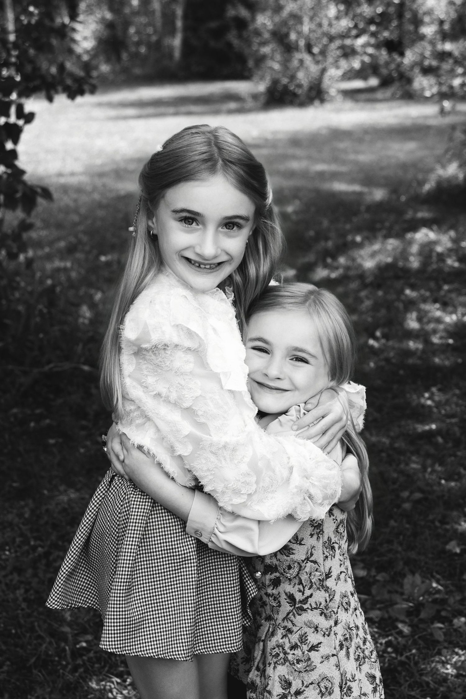 Two young girls hugging and smiling outdoors in a black and white portrait photo in a park setting.