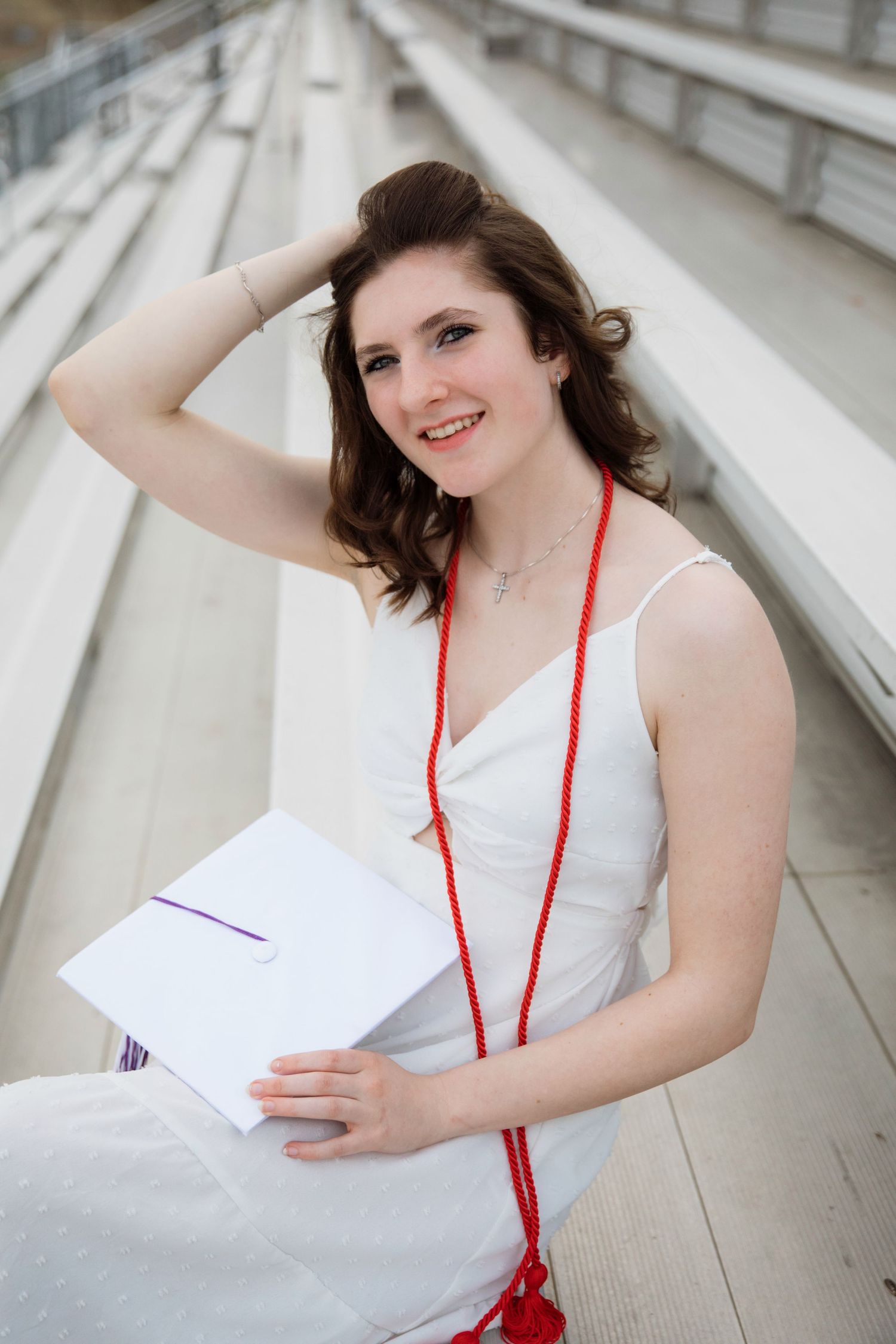 Smiling young woman in white dress with red graduation cord holds diploma while sitting in stadium bleachers.