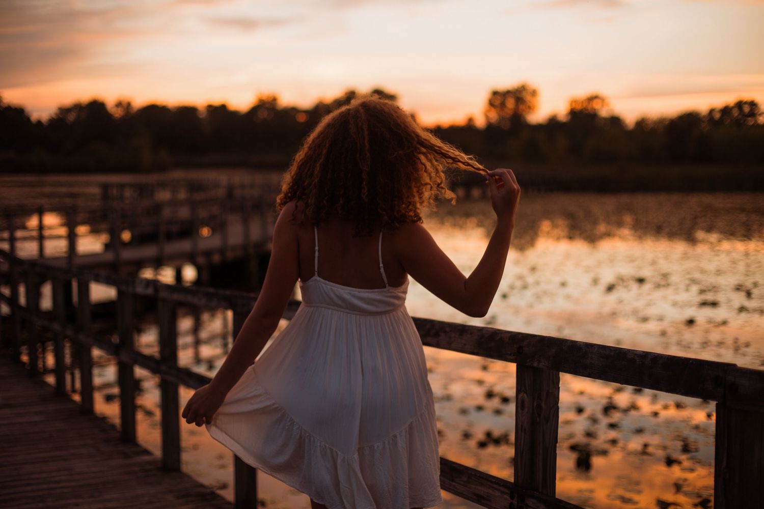 Woman in a white dress stands on a wooden bridge overlooking a lake at dramatic sunset.