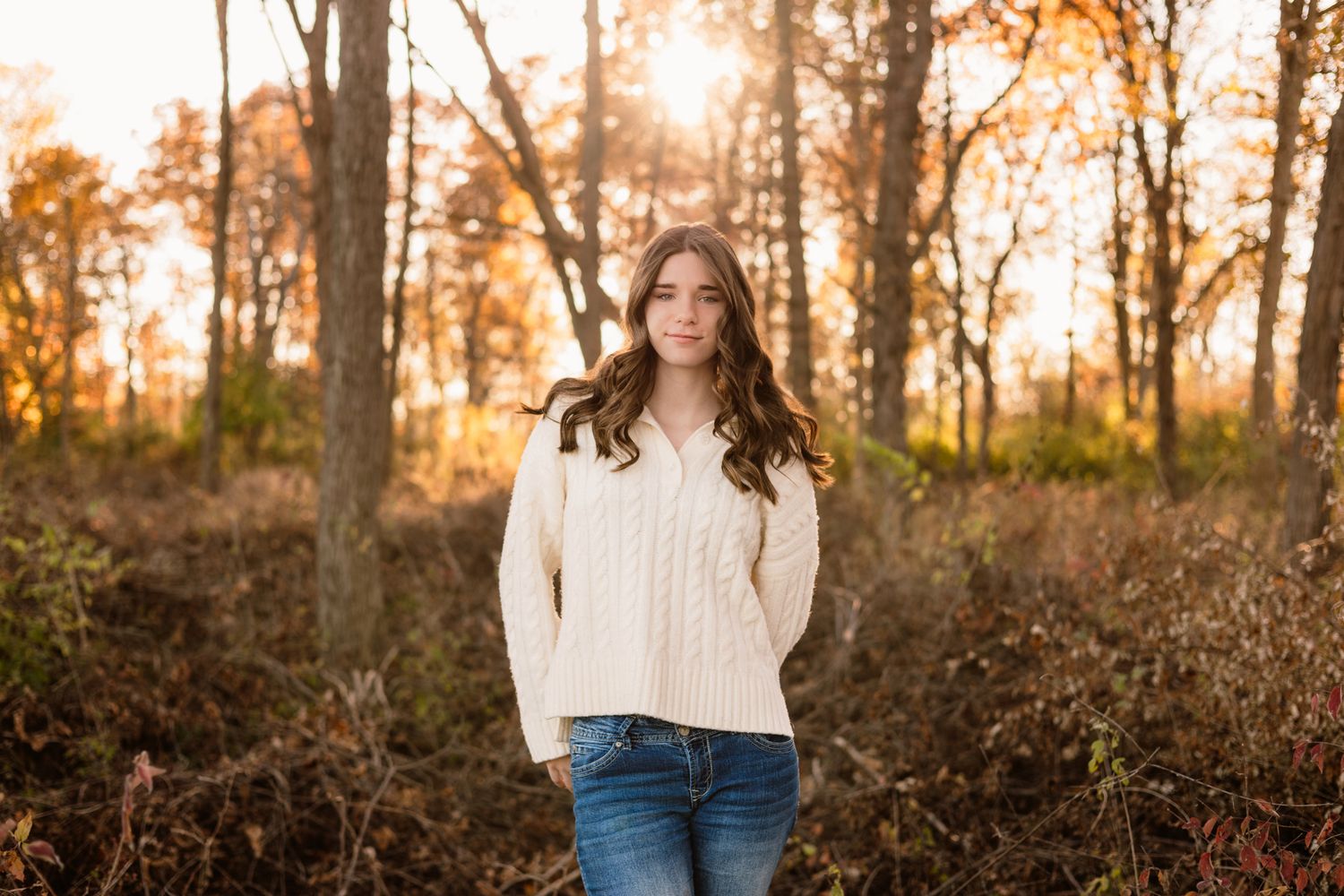 Young woman in cream sweater and jeans standing in autumn forest with golden sunset light behind her.