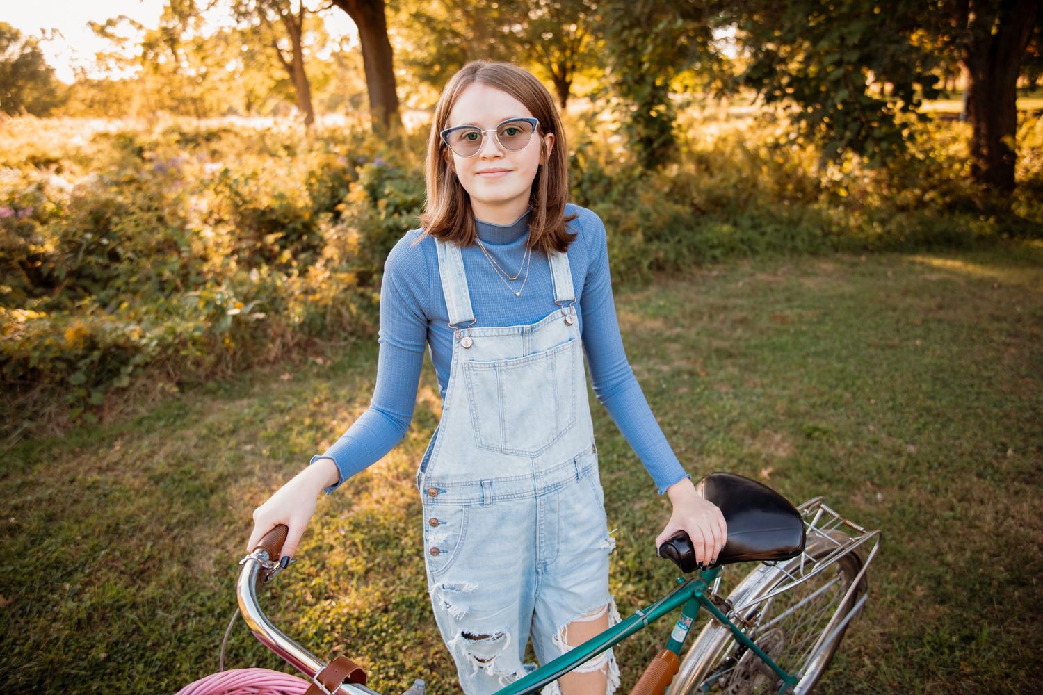 A young girl in denim overalls and sunglasses stands with a vintage bicycle in a sunny green park.
