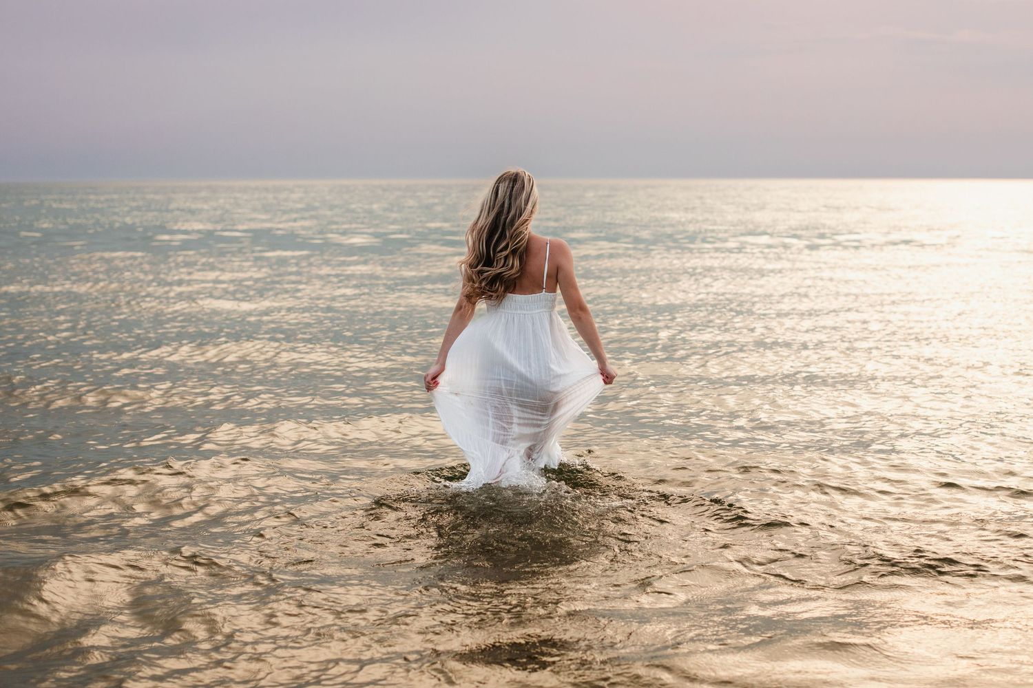 Woman in white dress wading in calm ocean water at sunset, viewed from behind.