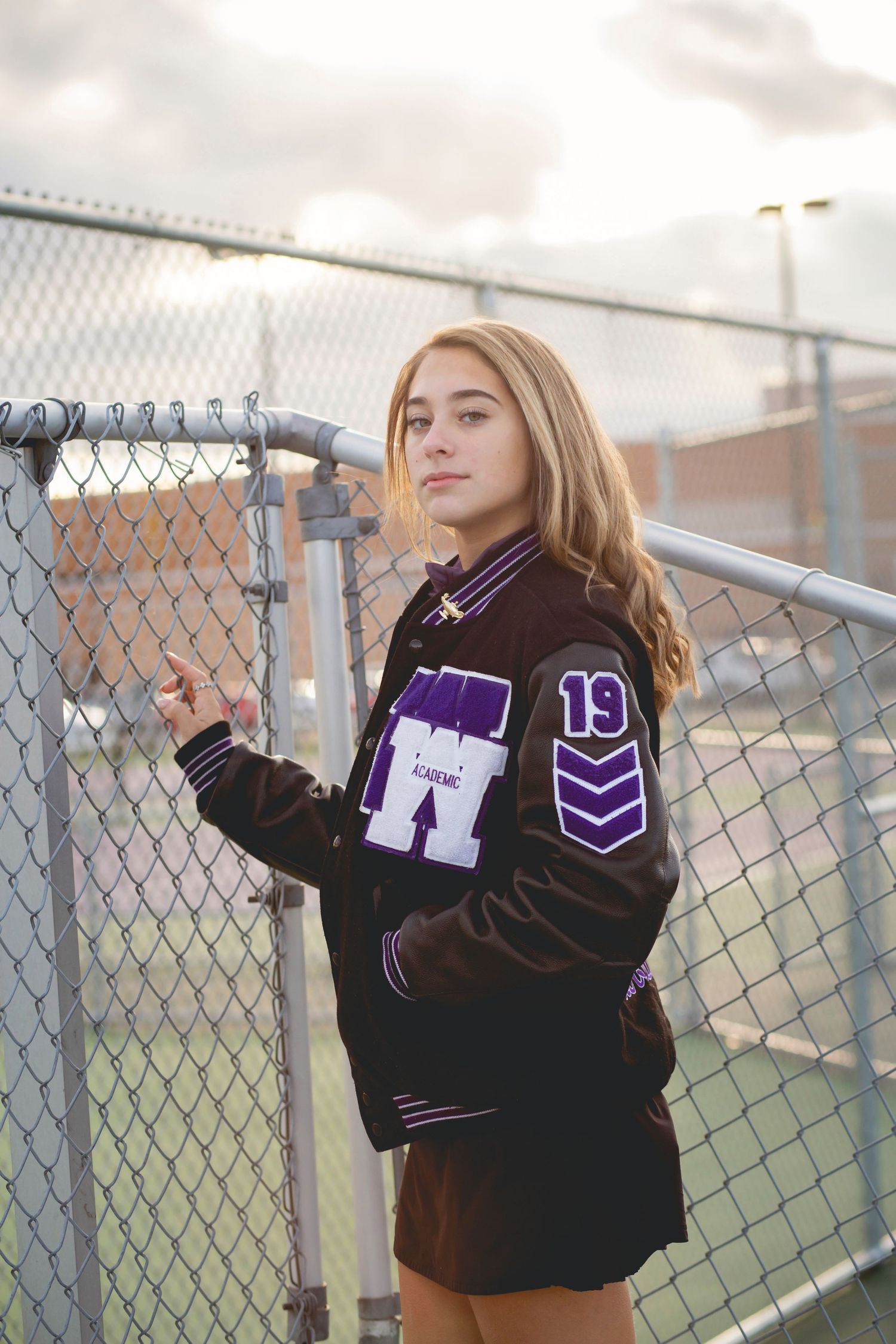 Teen girl in varsity letterman jacket with number 19 posing by chain-link fence at school athletic field.