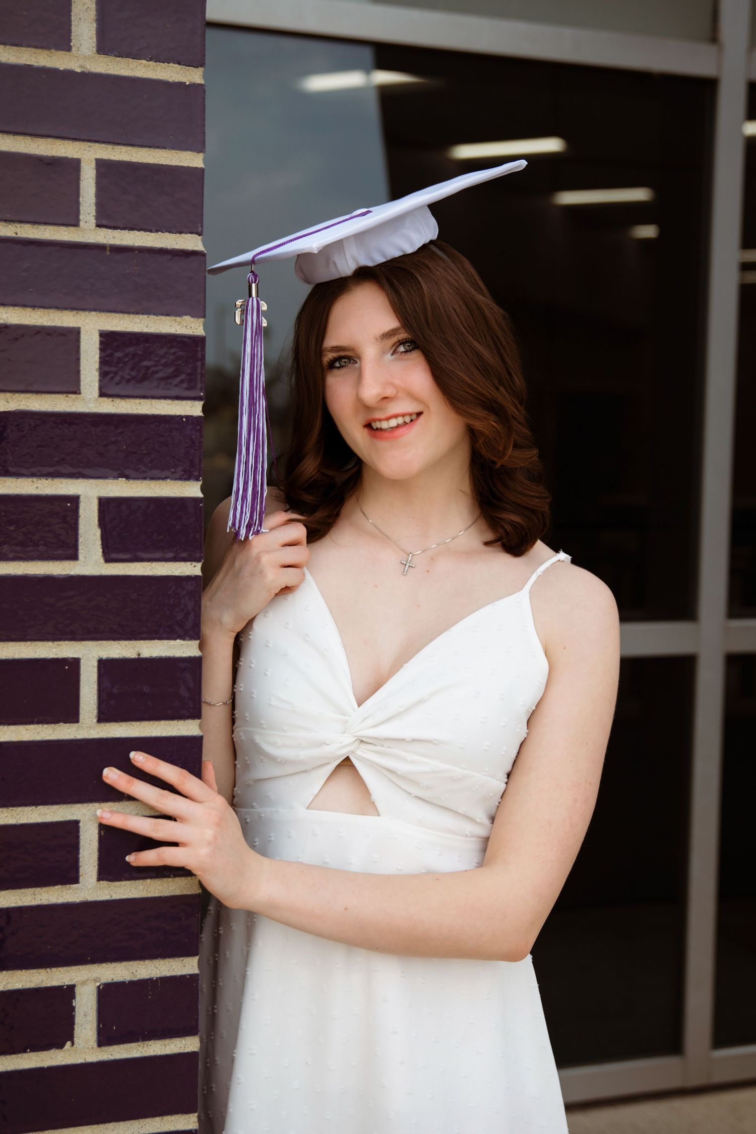 Young woman in white dress and purple graduation cap smiles proudly while leaning against a brick wall.