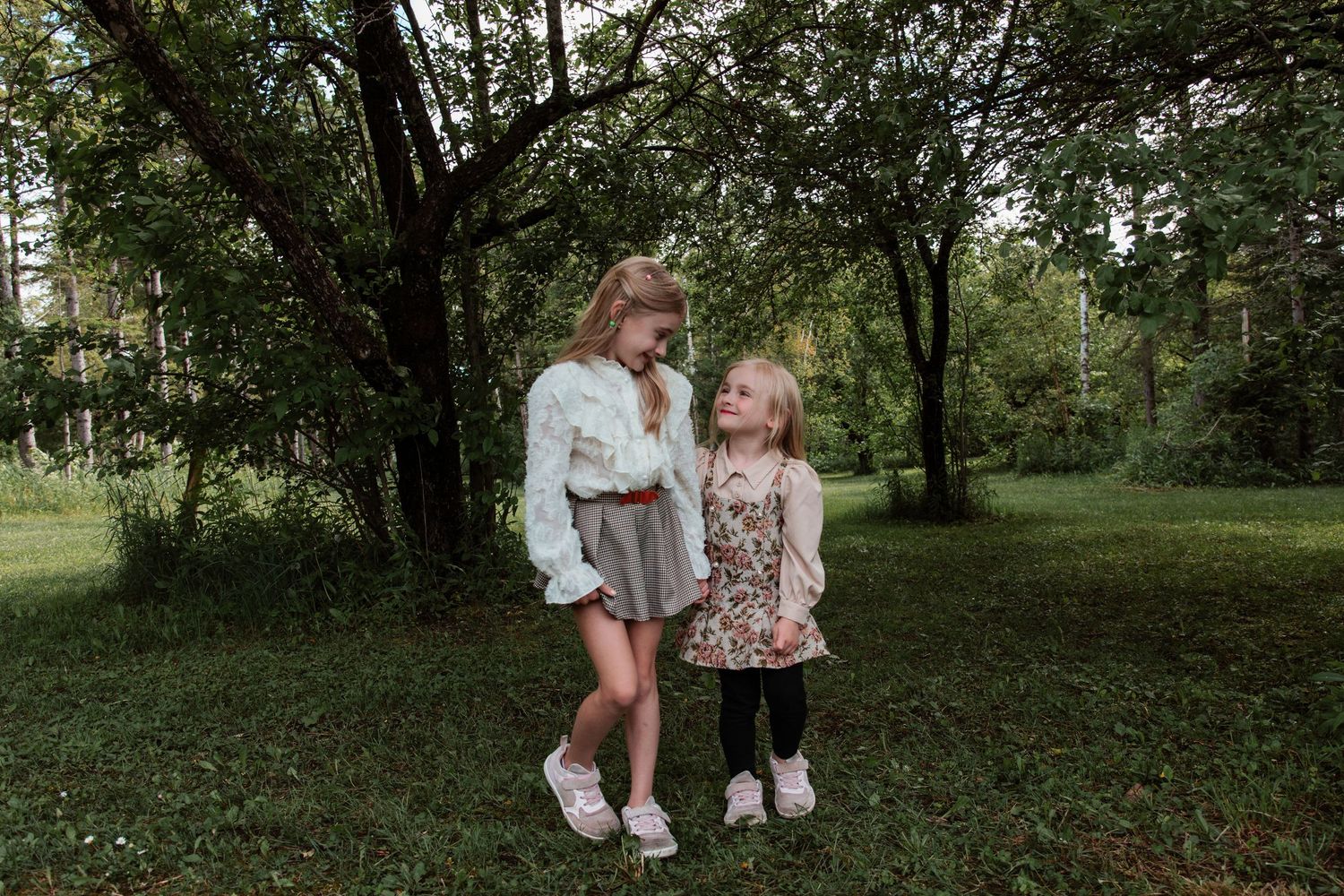 Two young girls in dresses smile at each other while standing on green grass surrounded by trees.