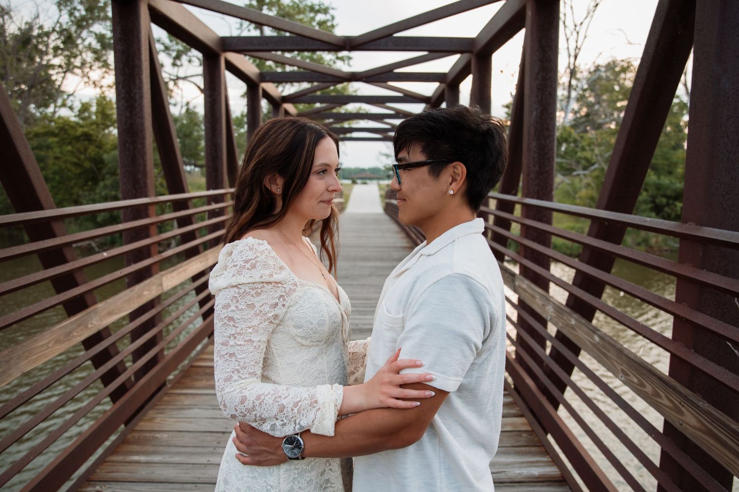 Couple in white outfits embracing and gazing at each other on a rustic wooden bridge outdoors.