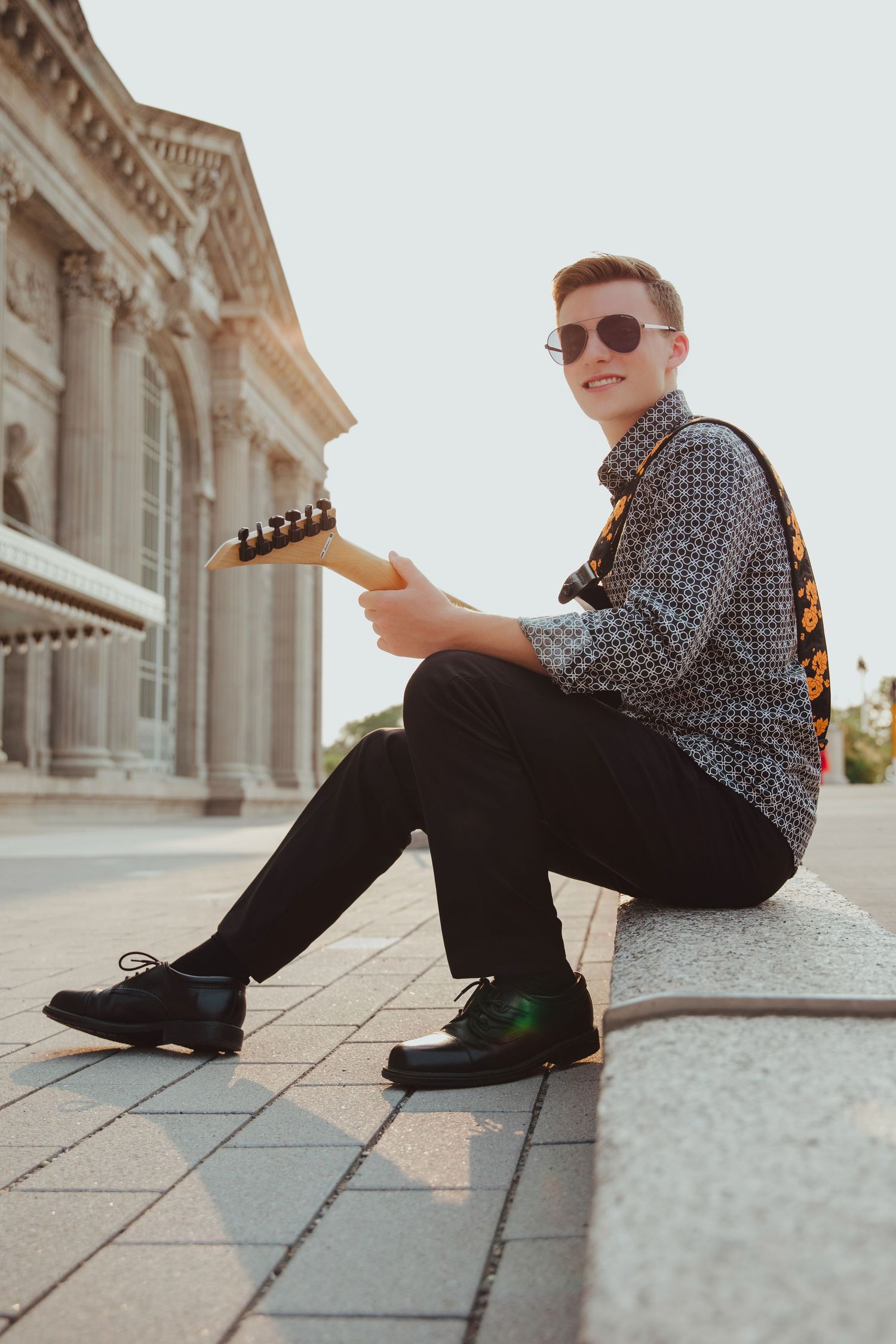 Smiling young man in sunglasses sitting outdoors holding electric guitar.