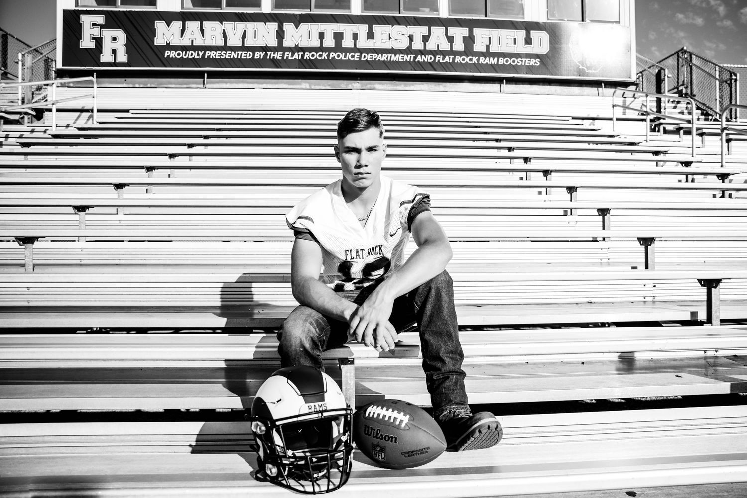 Black and white photo of a young male athlete posing with football helmet and ball in stadium bleachers at Marvin Mittlestat Field.
