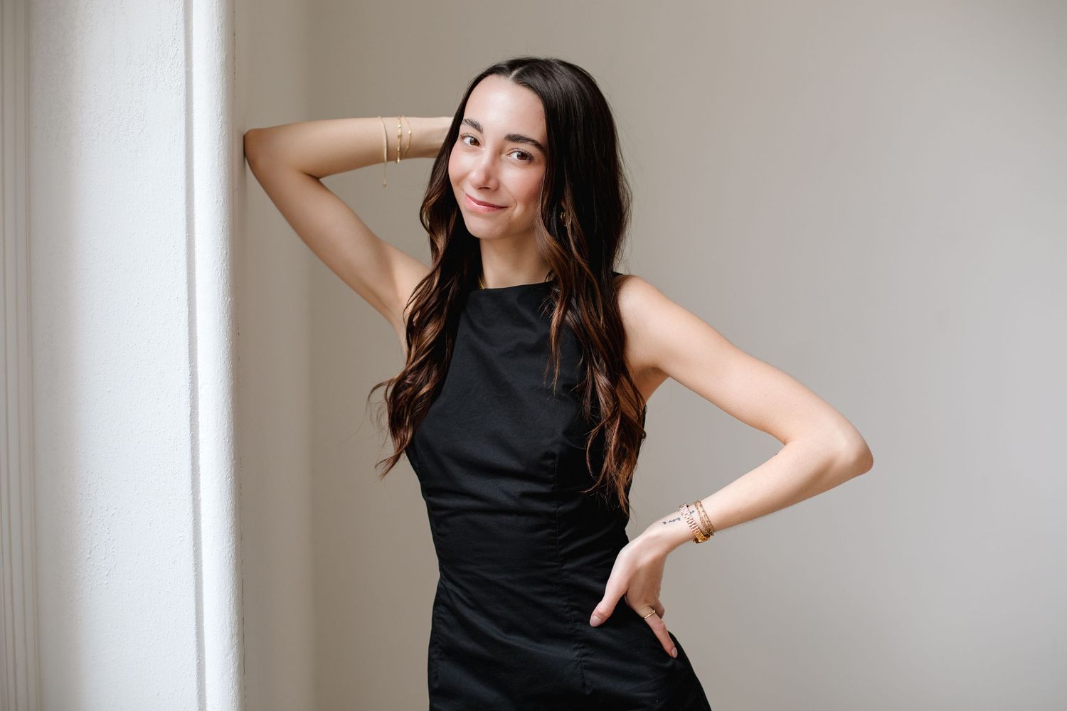 Young woman in black sleeveless dress posing confidently against light wall with hand on hip.