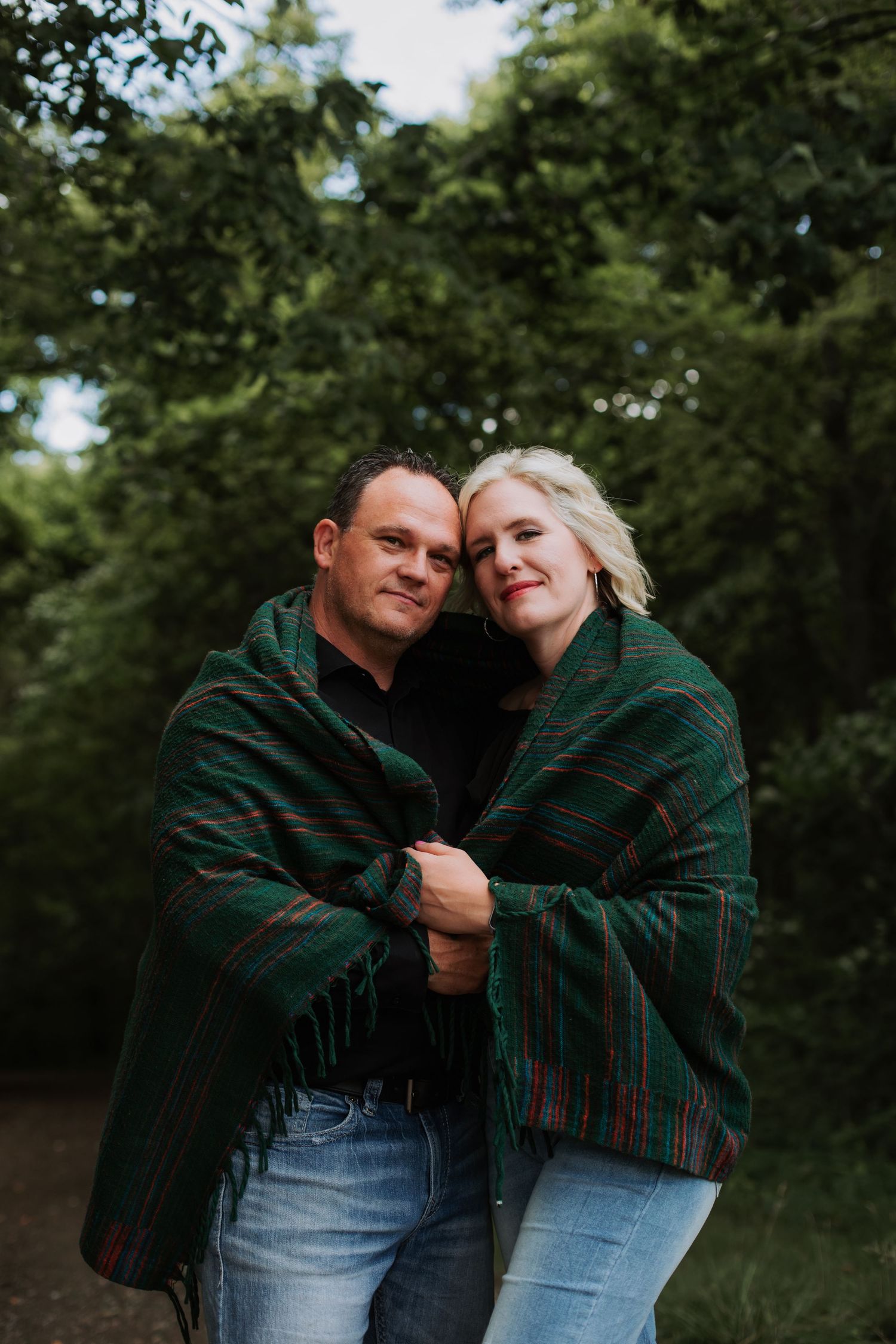 Couple wrapped in a plaid blanket sharing a cozy forest portrait.