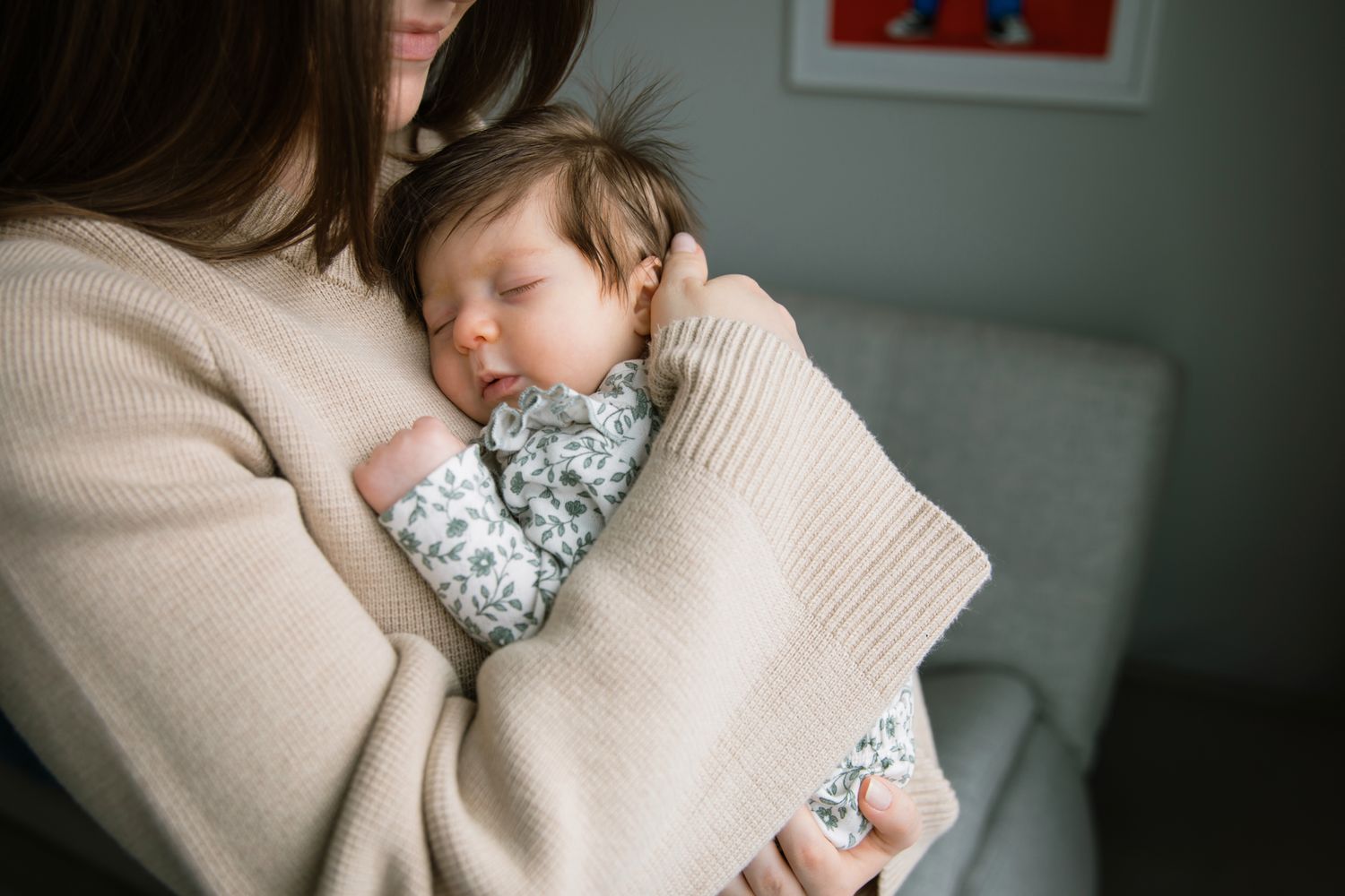 Mother in beige sweater cradling sleeping newborn baby against her chest.