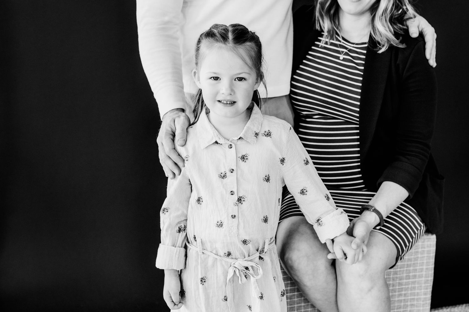 Young girl stands smiling in front of her seated pregnant mother and standing father in black and white.
