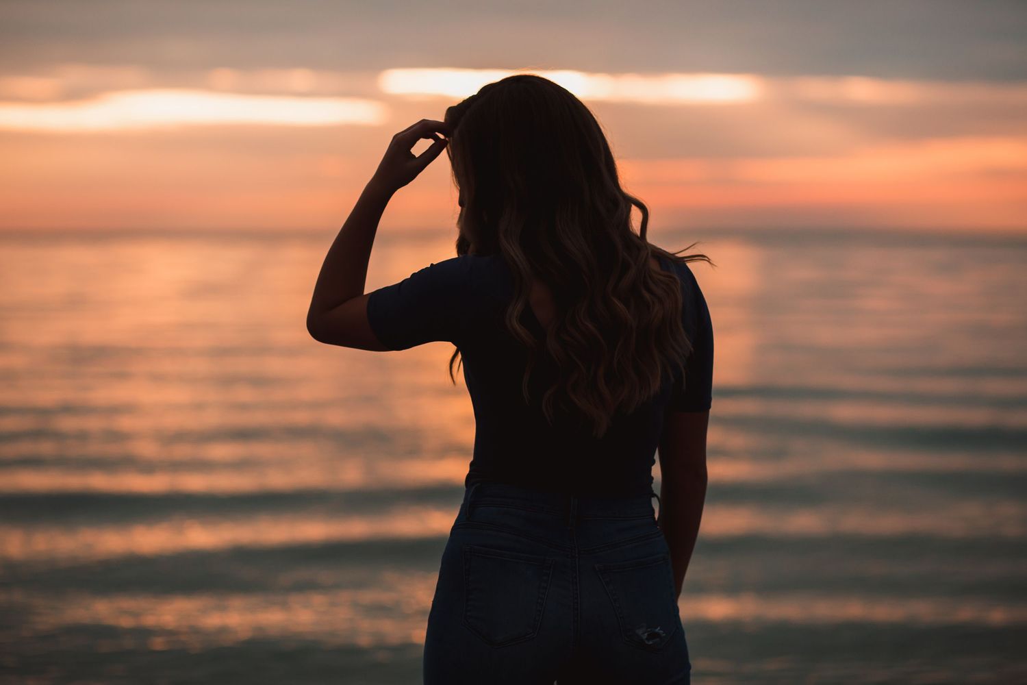 Silhouette of a young woman with long wavy hair standing at the ocean shore during a vibrant sunset.