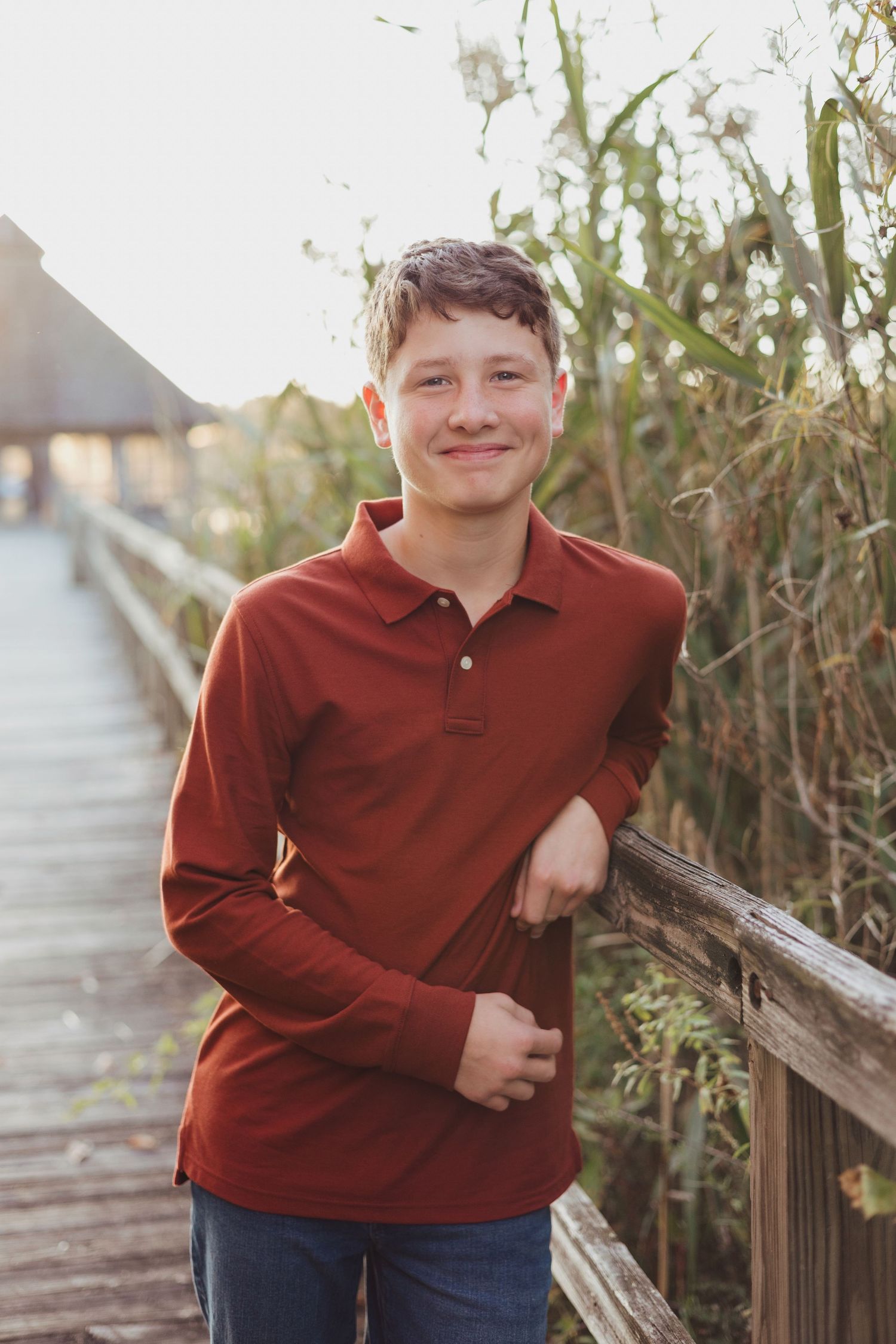 Teenage boy in red polo shirt leaning on wooden railing on a boardwalk surrounded by tall grasses.