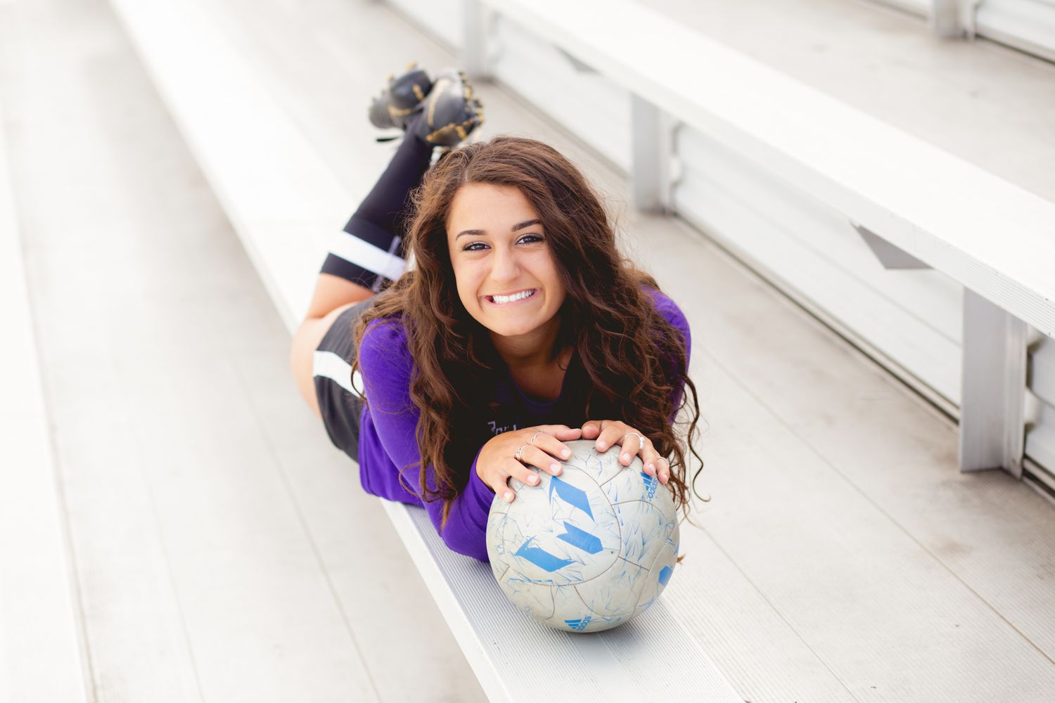 Smiling teenage girl in purple jersey poses with soccer ball on bleachers for senior photos.