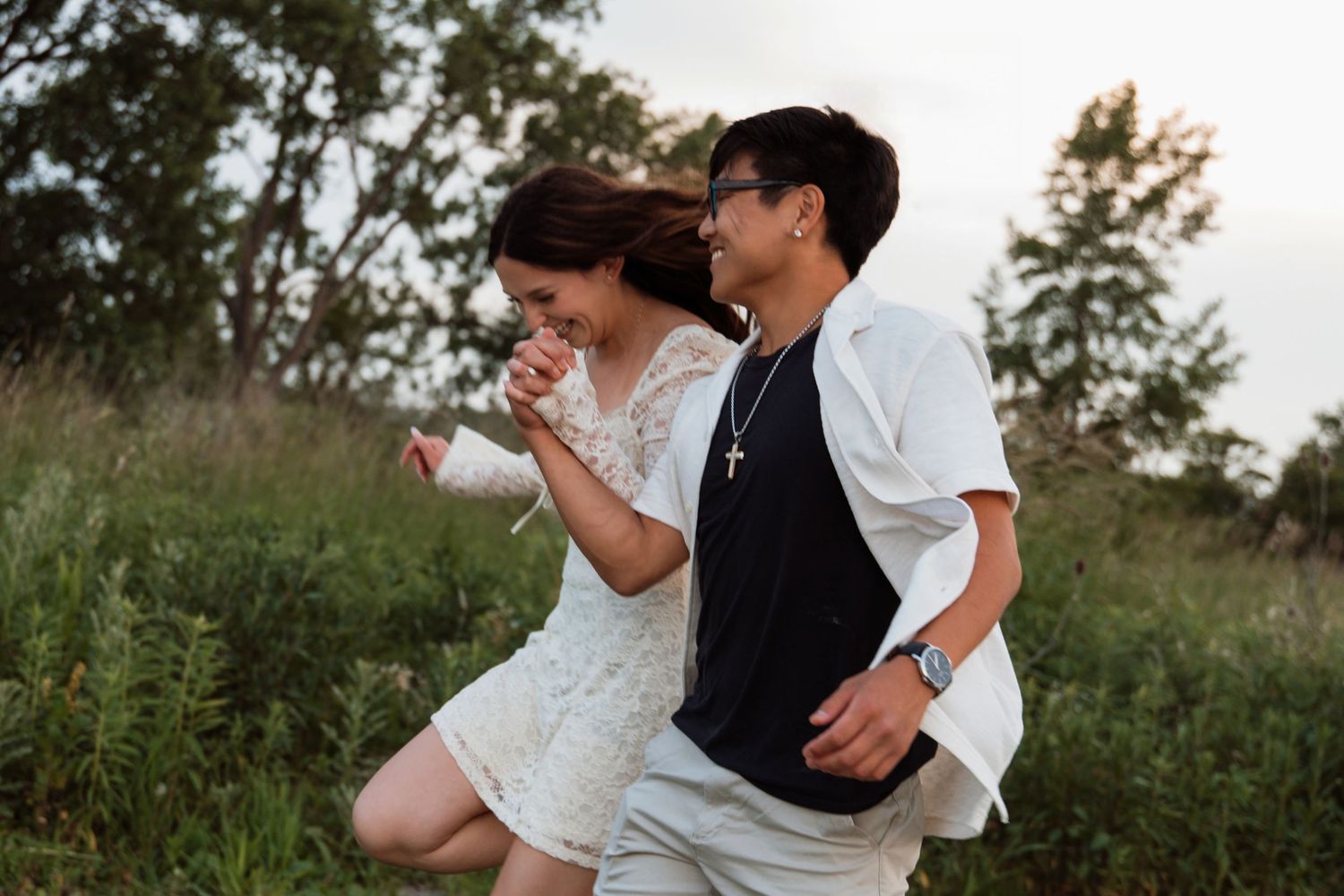 Playful couple running and laughing together through a green field at sunset.