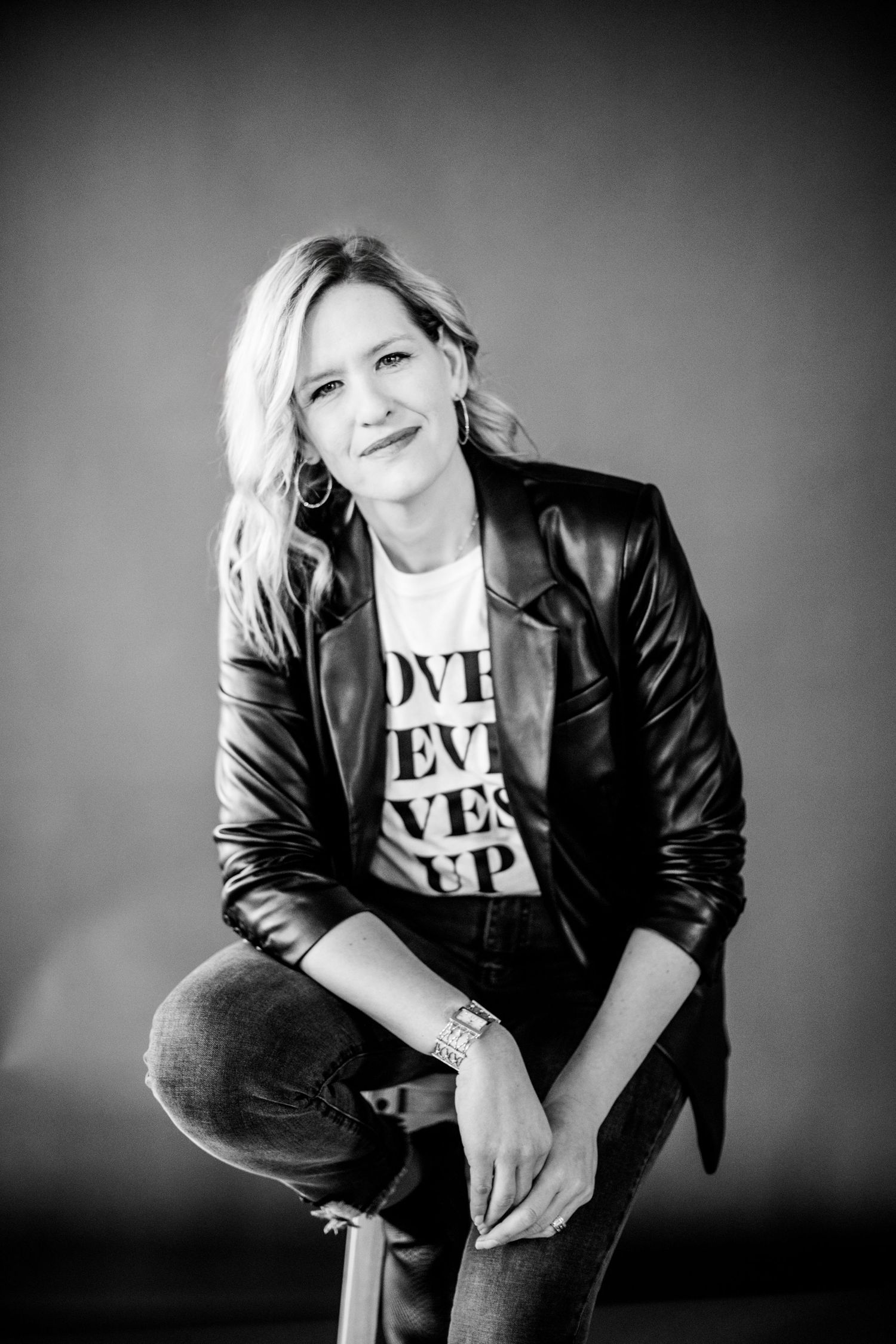 Black and white portrait of a blonde woman in a leather jacket seated on a stool smiling.