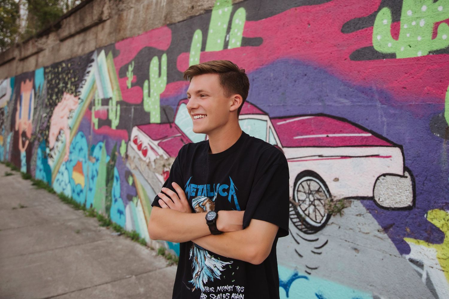 A smiling young man in a black t-shirt standing in front of a colorful graffiti mural with arms crossed.