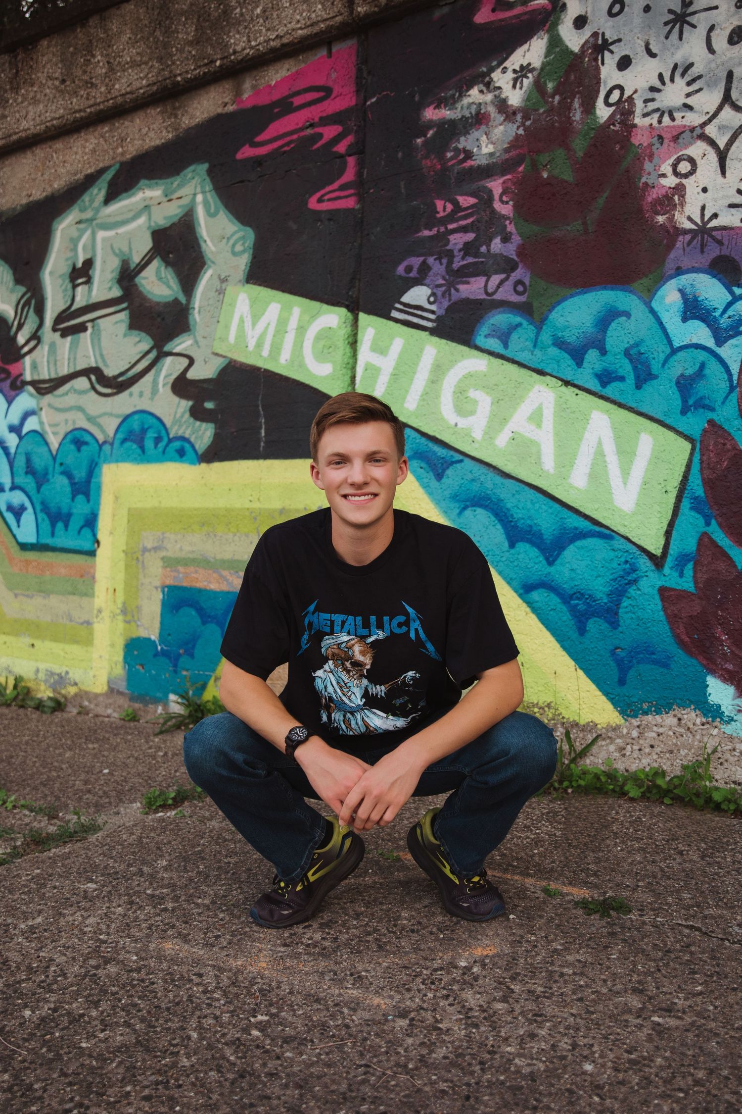 Smiling teen crouches before Michigan graffiti mural wearing Metallica shirt and jeans.
