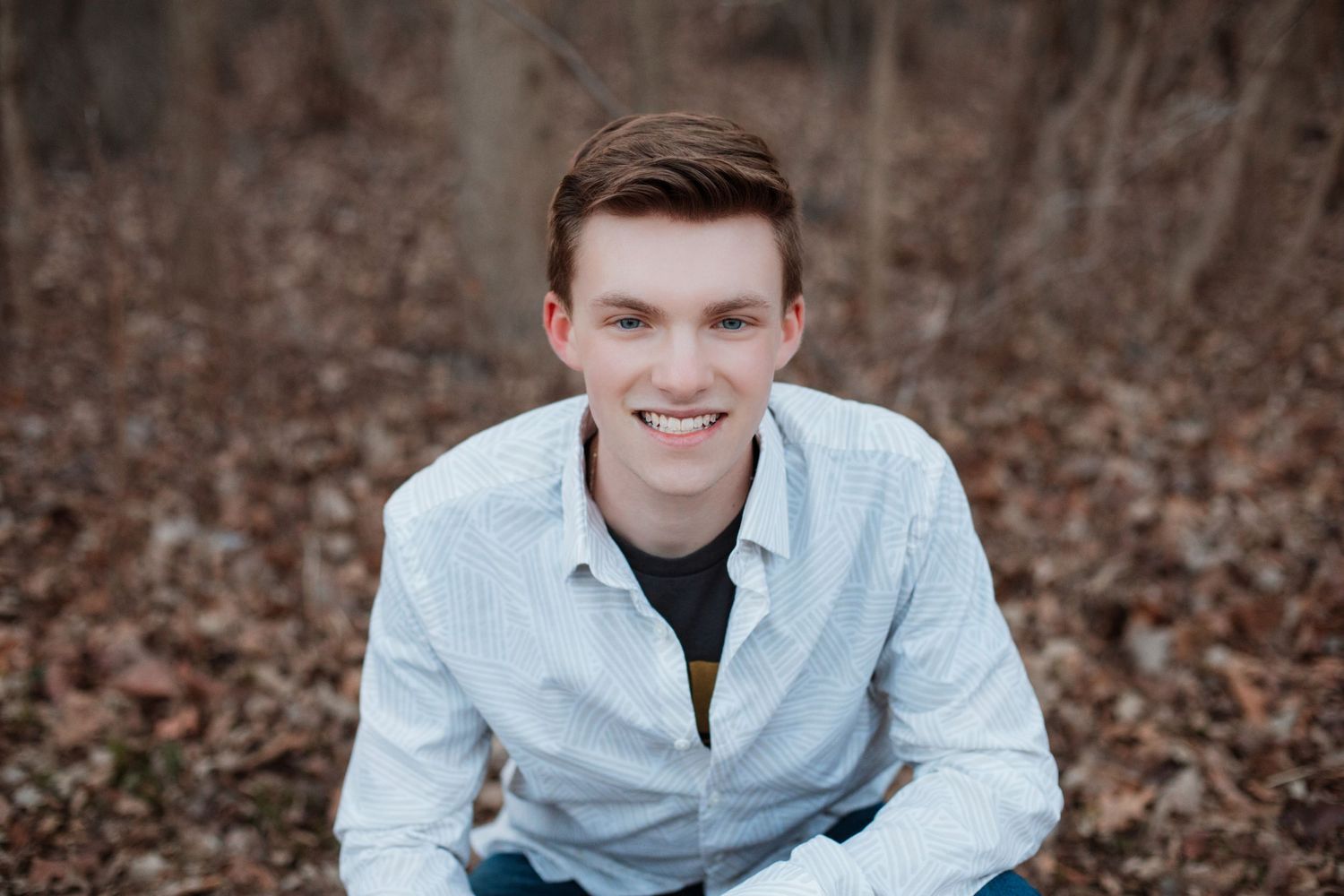 Smiling young man in light blue shirt sits outdoors among autumn leaves in a natural setting.