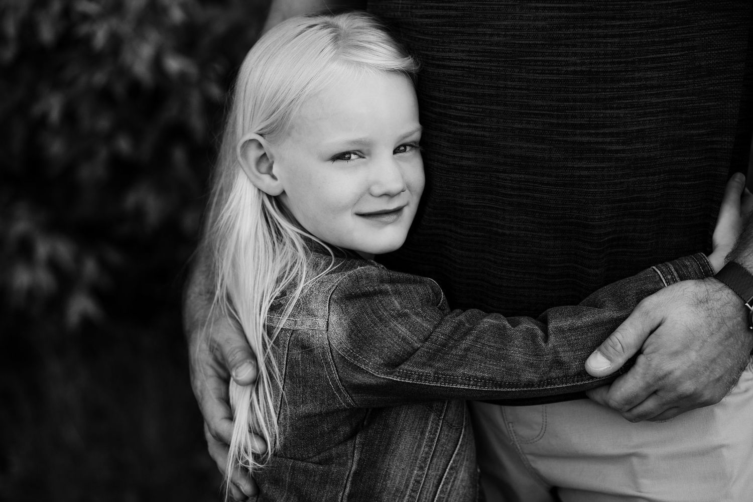 Young blonde girl smiling while hugging adult outdoors in black and white photo.