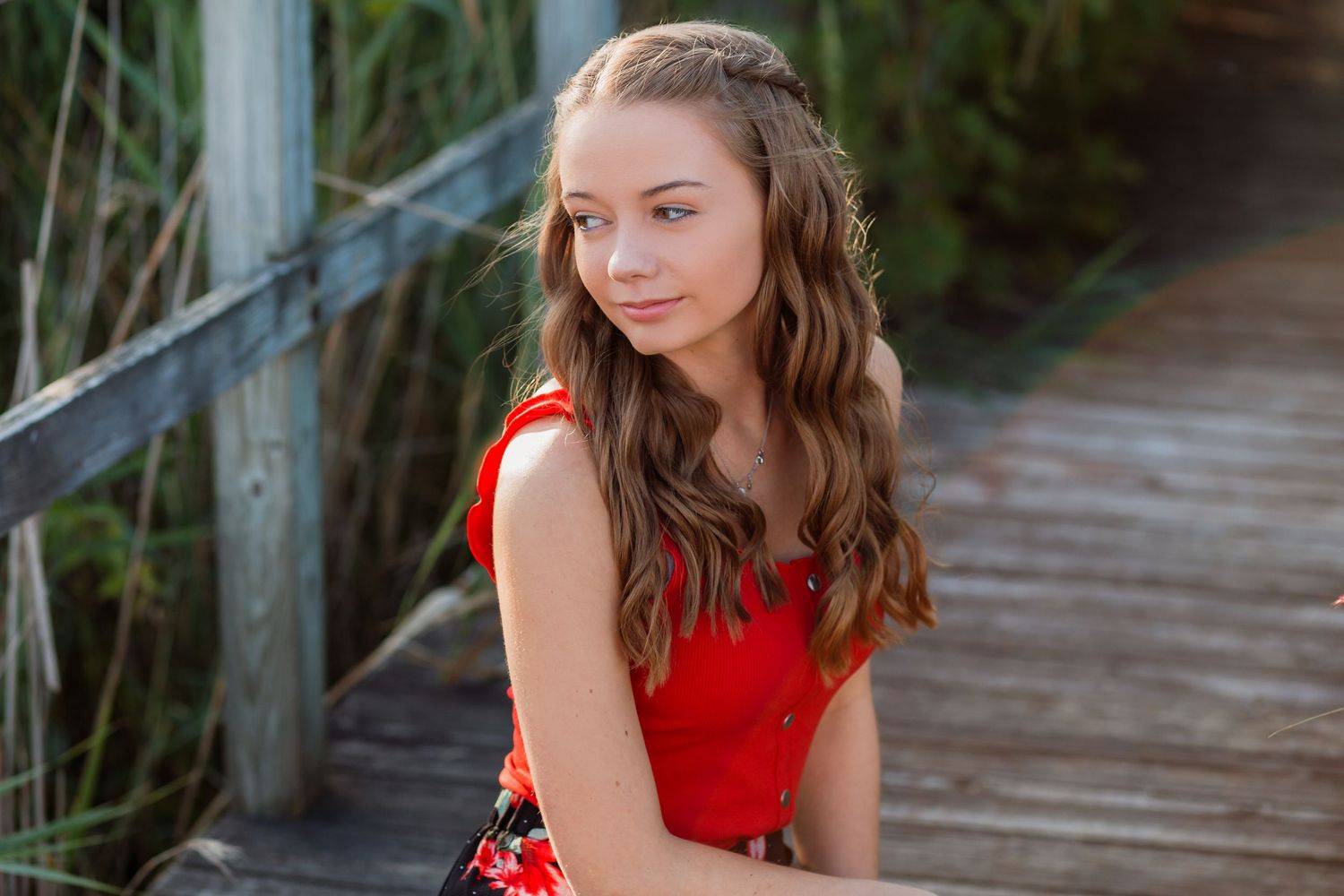 Young girl in red floral dress sitting on wooden boardwalk surrounded by tall grass.