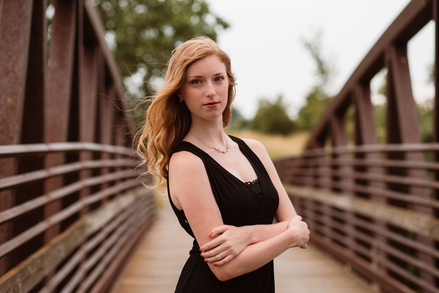 Young redhead woman in black dress standing confidently on a rustic bridge outdoors.