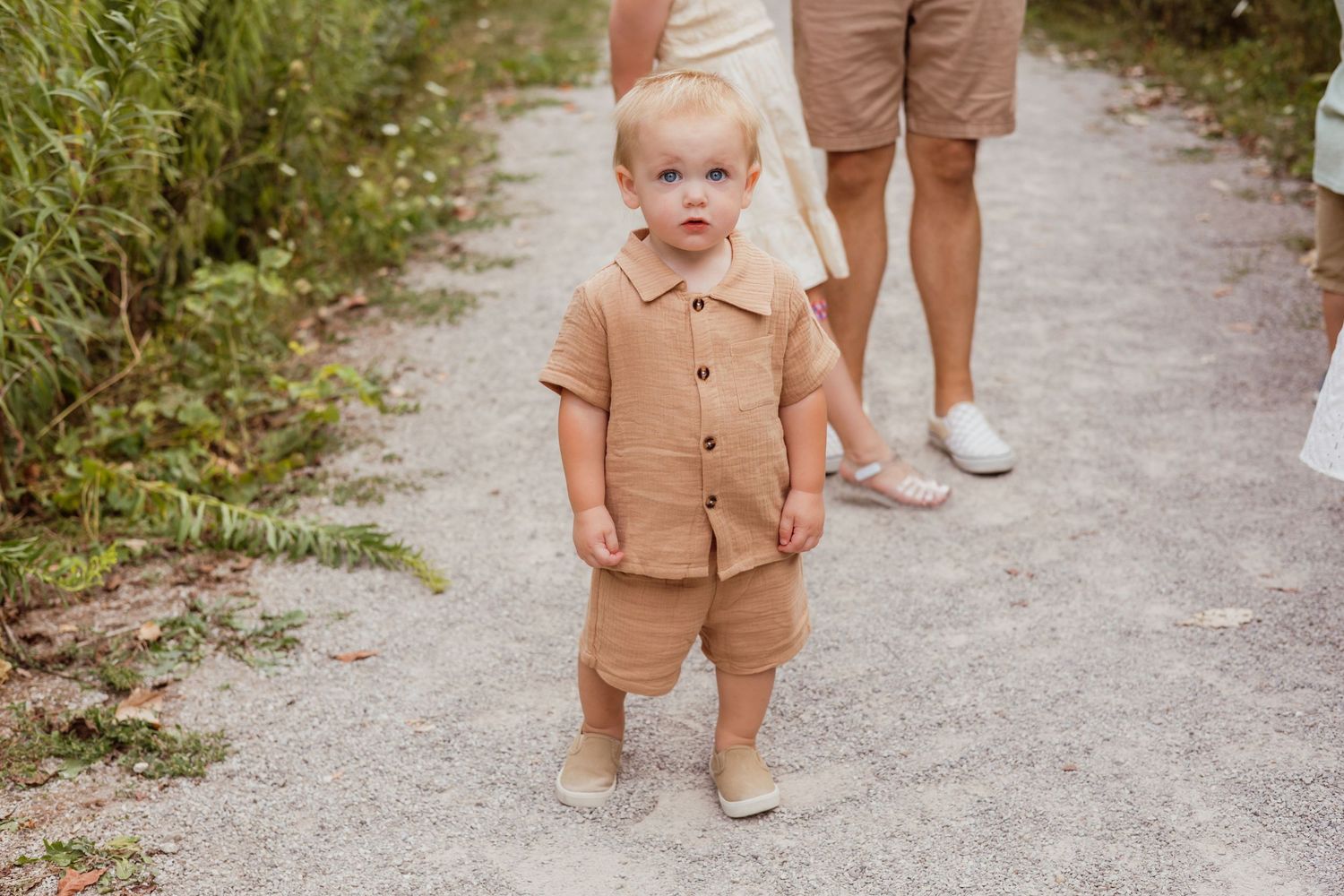 A toddler in a tan linen outfit stands on a gravel path outdoors.