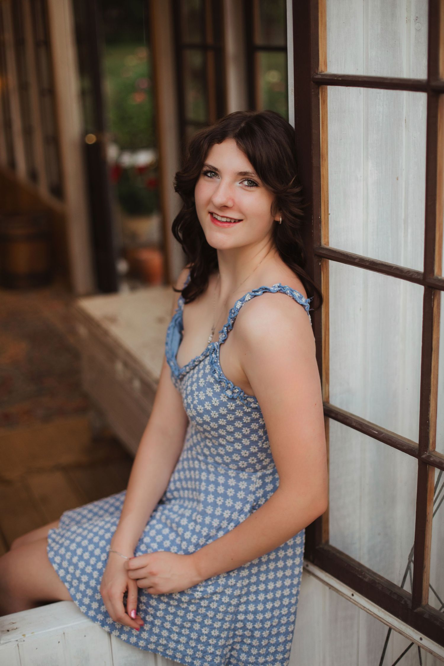 Young woman in blue polka dot dress sitting and smiling by a white wooden door frame.