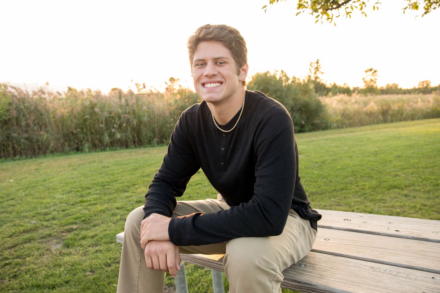 Smiling young man in black shirt sits on wooden bench outdoors at golden hour with green grass and reeds behind him.
