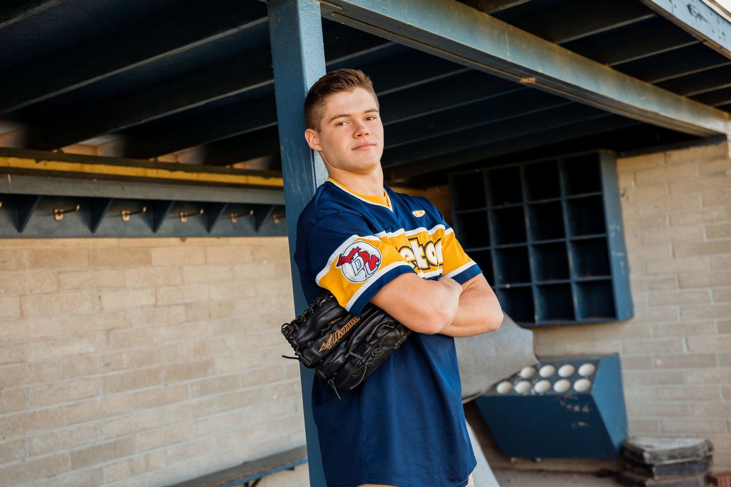 Young baseball player in navy uniform holding glove, posing confidently in dugout at stadium.